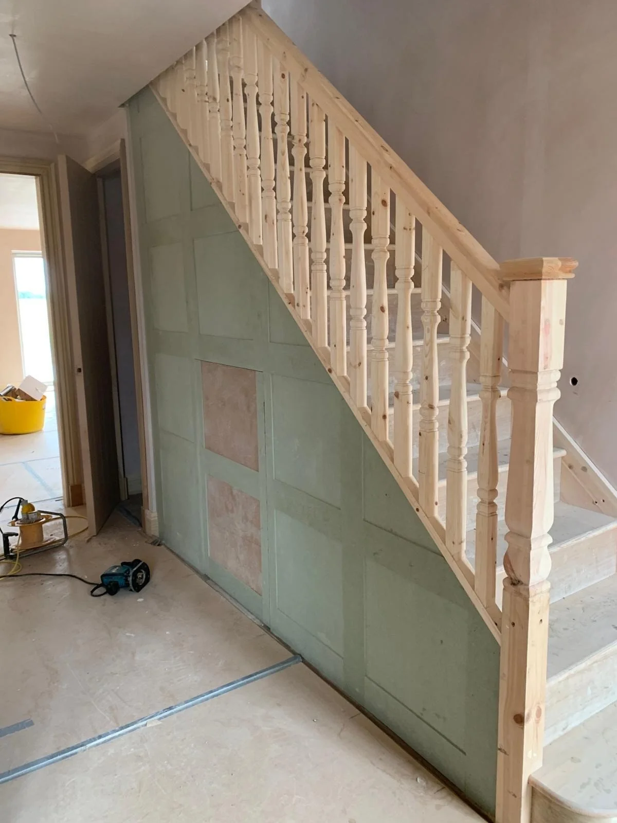 Wooden staircase with balusters and newel post under construction in a house interior, with unfinished walls and construction tools on the floor.