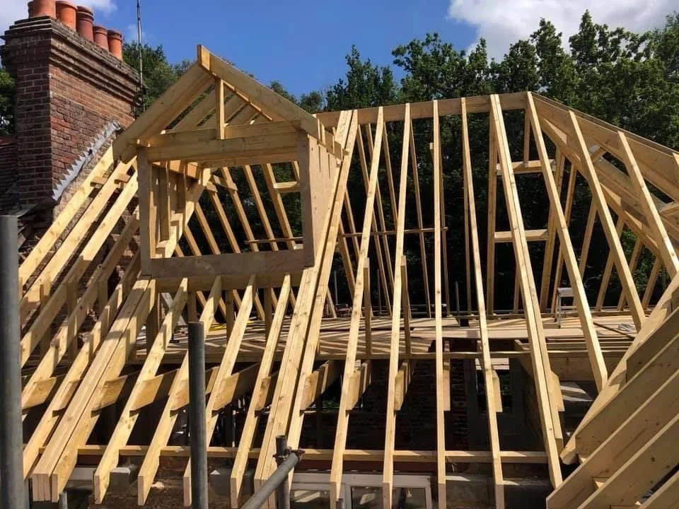 Wooden roof framing for a house under construction, with a chimney on a brick wall in the background and a clear sky.