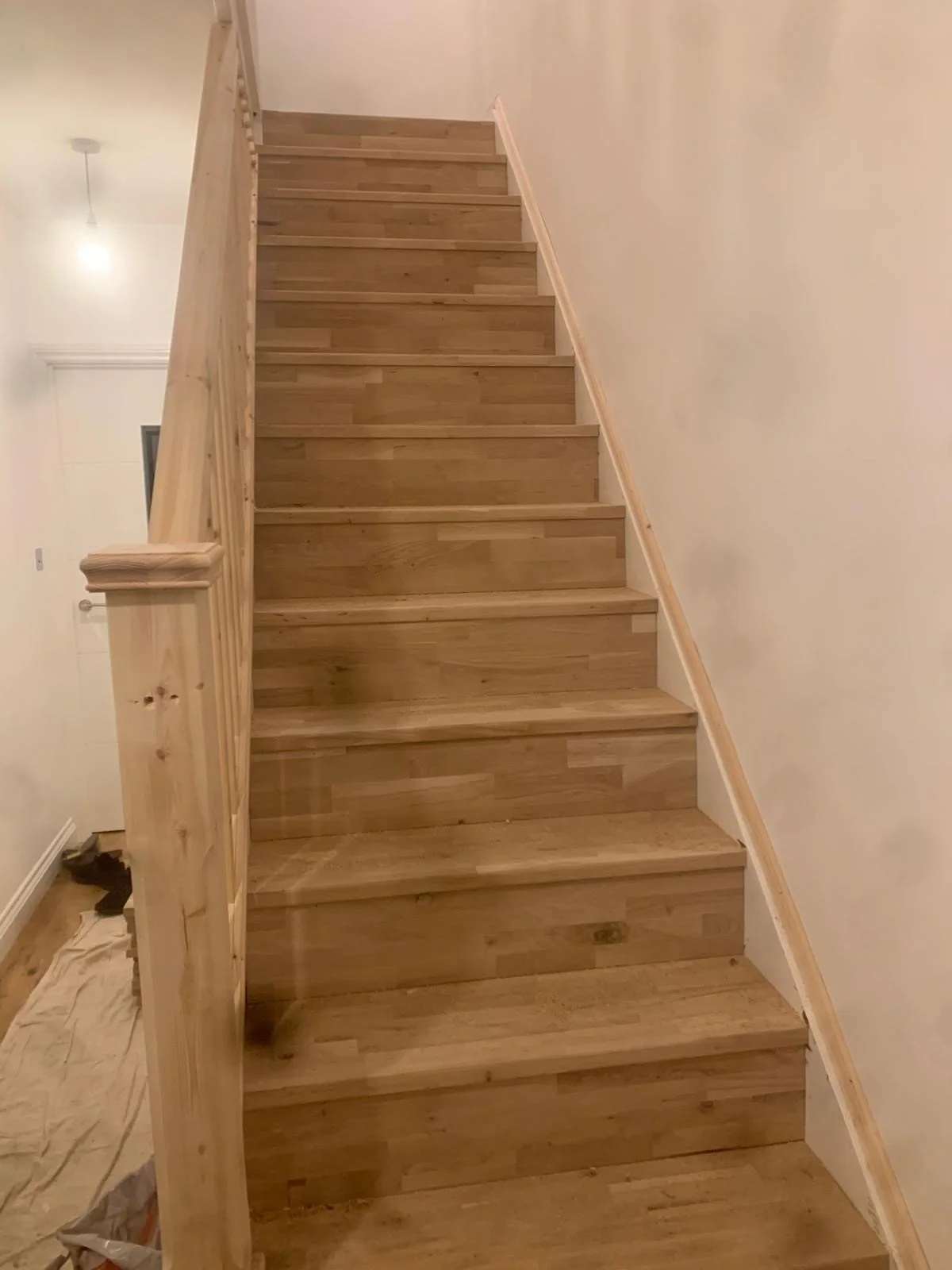 Wood staircase with a newel post on the left and a handrail on the right, leading upwards along a white wall in a home interior.