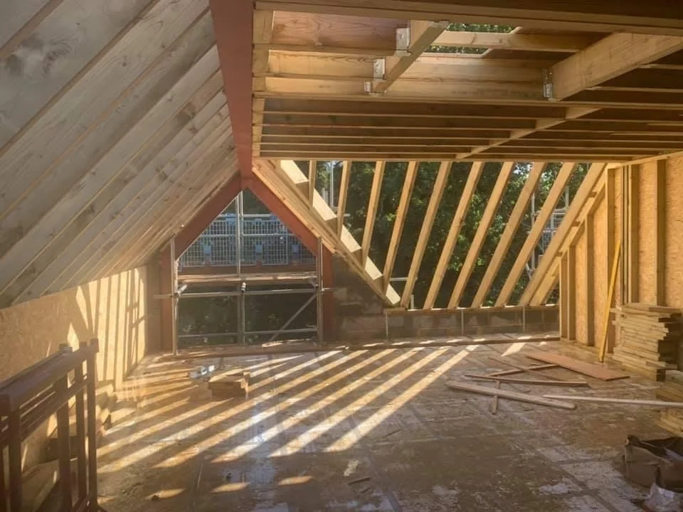 Interior of a building under construction with exposed wooden framing and structural supports, sunlight casting shadows through the open framework, scattered construction materials on the floor.