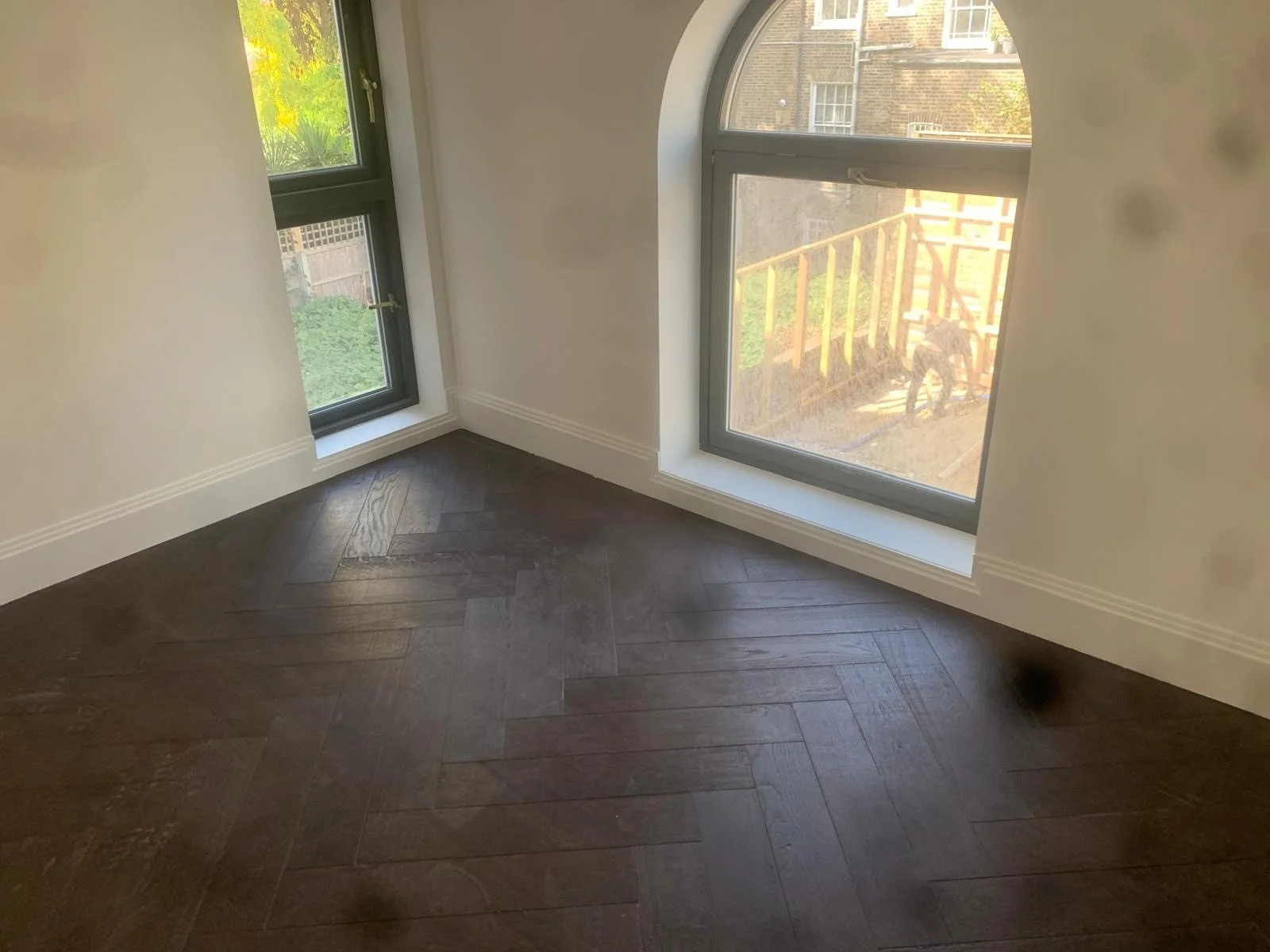 Empty room with dark wooden floor and white walls, featuring large windows with black frames allowing natural light, and an outdoor view of a backyard with a deck and a dog.