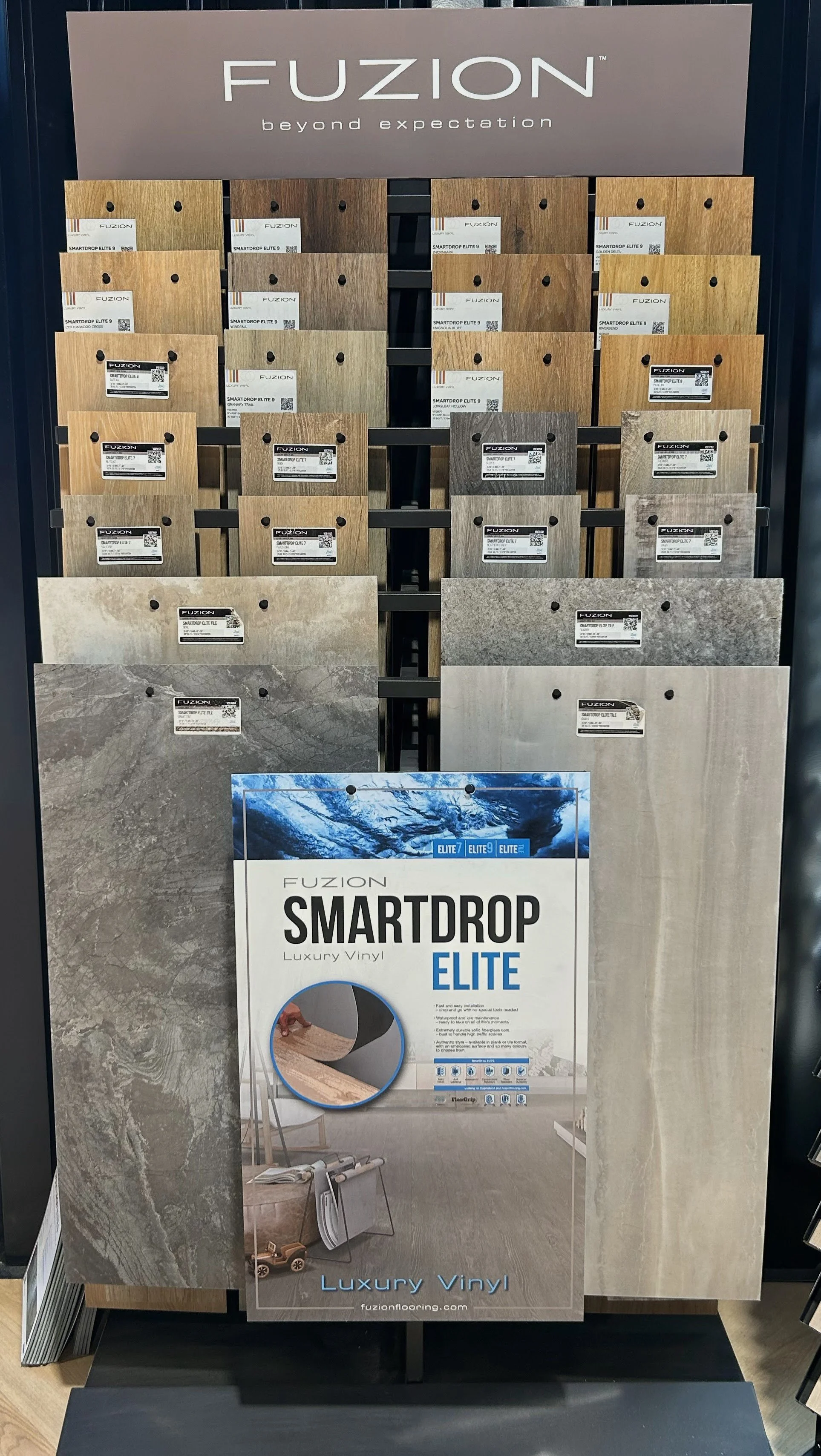 Display of Fuzion SmartDrop Elite luxury vinyl flooring samples in various wood and stone patterns, with a sign reading 'Fuzion beyond expectation' at the top.