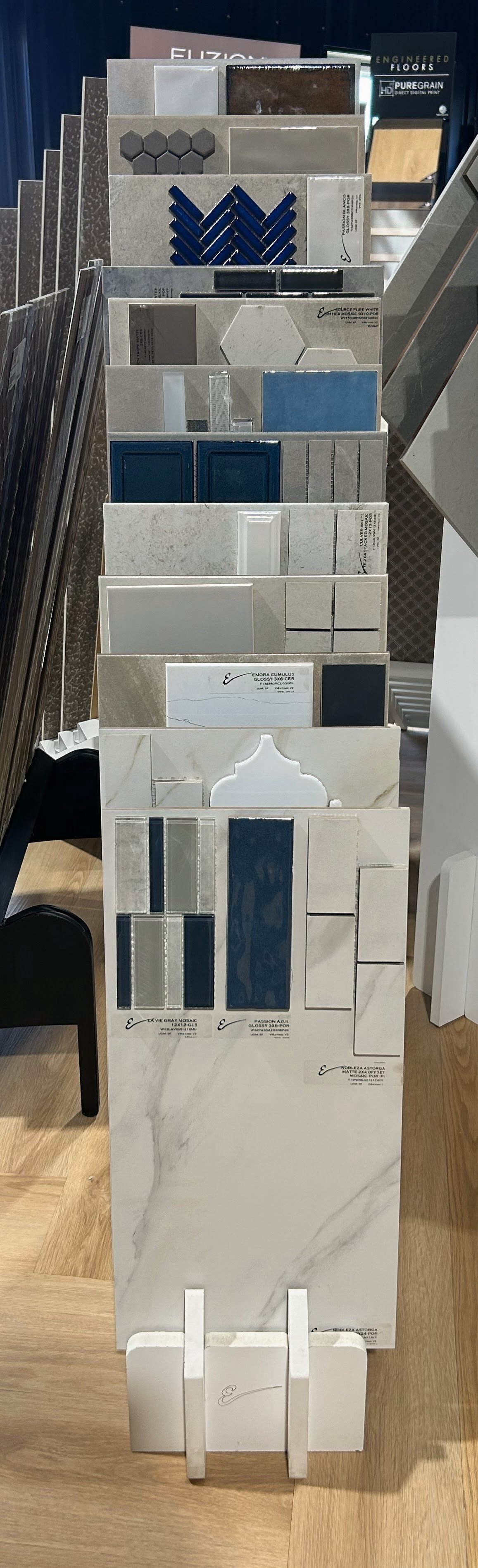 Display stand with various tile samples in shades of white, gray, blue, and black arranged vertically, some with patterns and textures, in a flooring or tile store.
