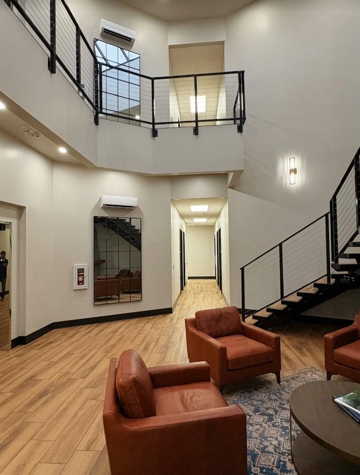 Interior of a modern building lobby with beige walls, wood flooring, leather chairs, a staircase, and upper-level balcony.