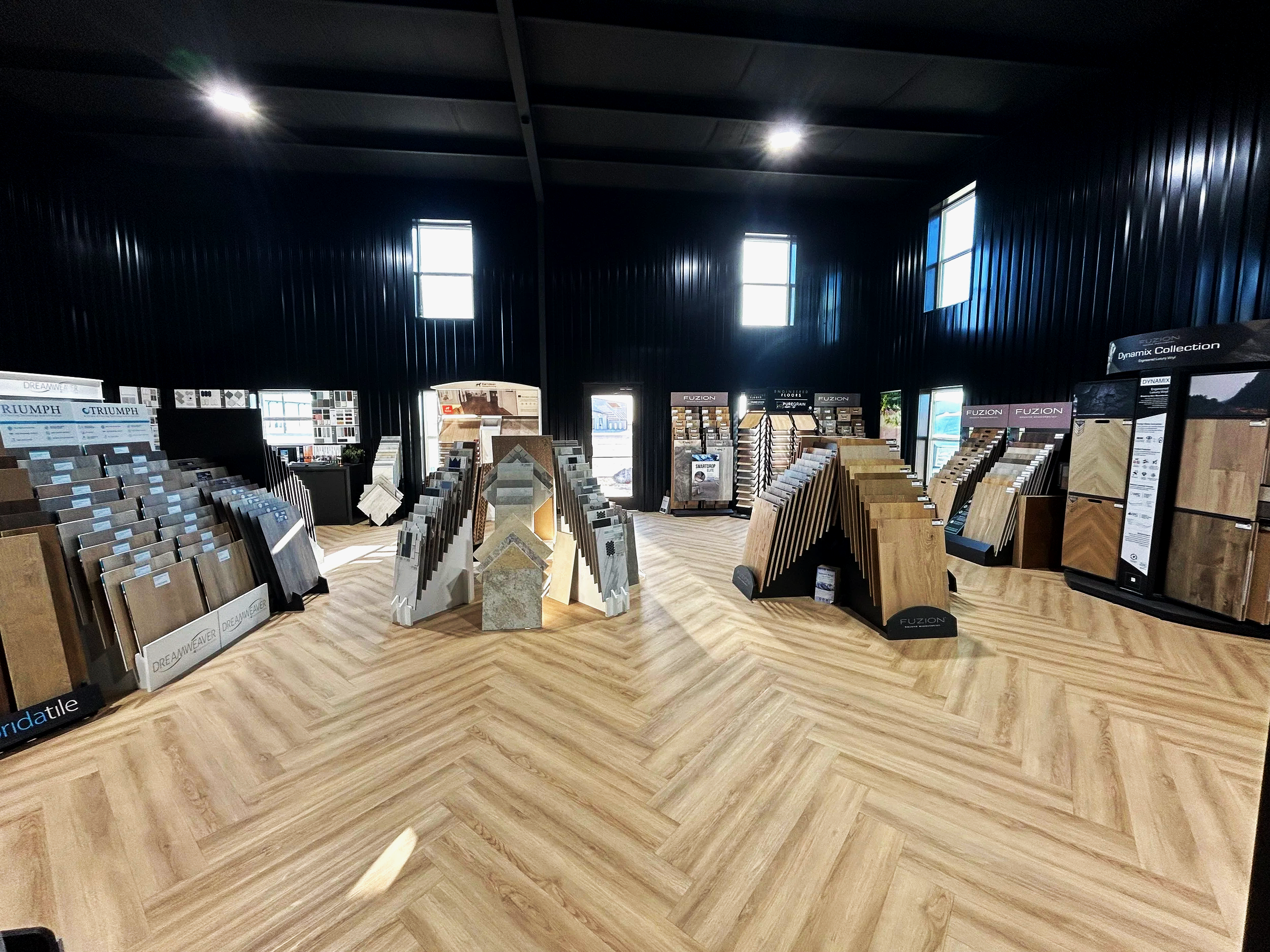Interior of a flooring showroom with sample display racks of hardwood and laminate flooring options arranged in diagonal rows on a light wood-patterned floor. Black walls and multiple windows illuminate the space.