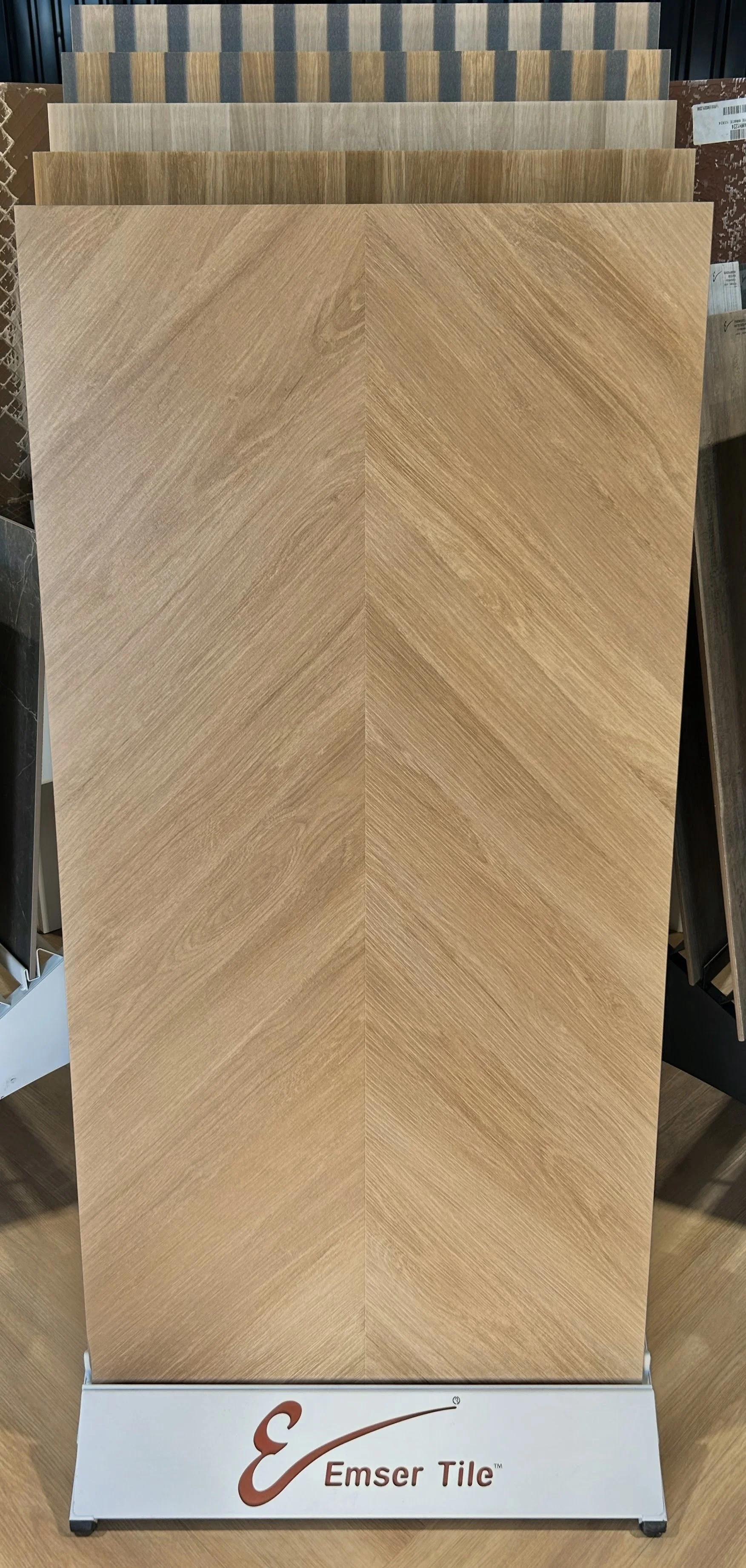 Wood grain tile samples displayed on a stand labeled Emser Tile, with various shades of brown and tan.