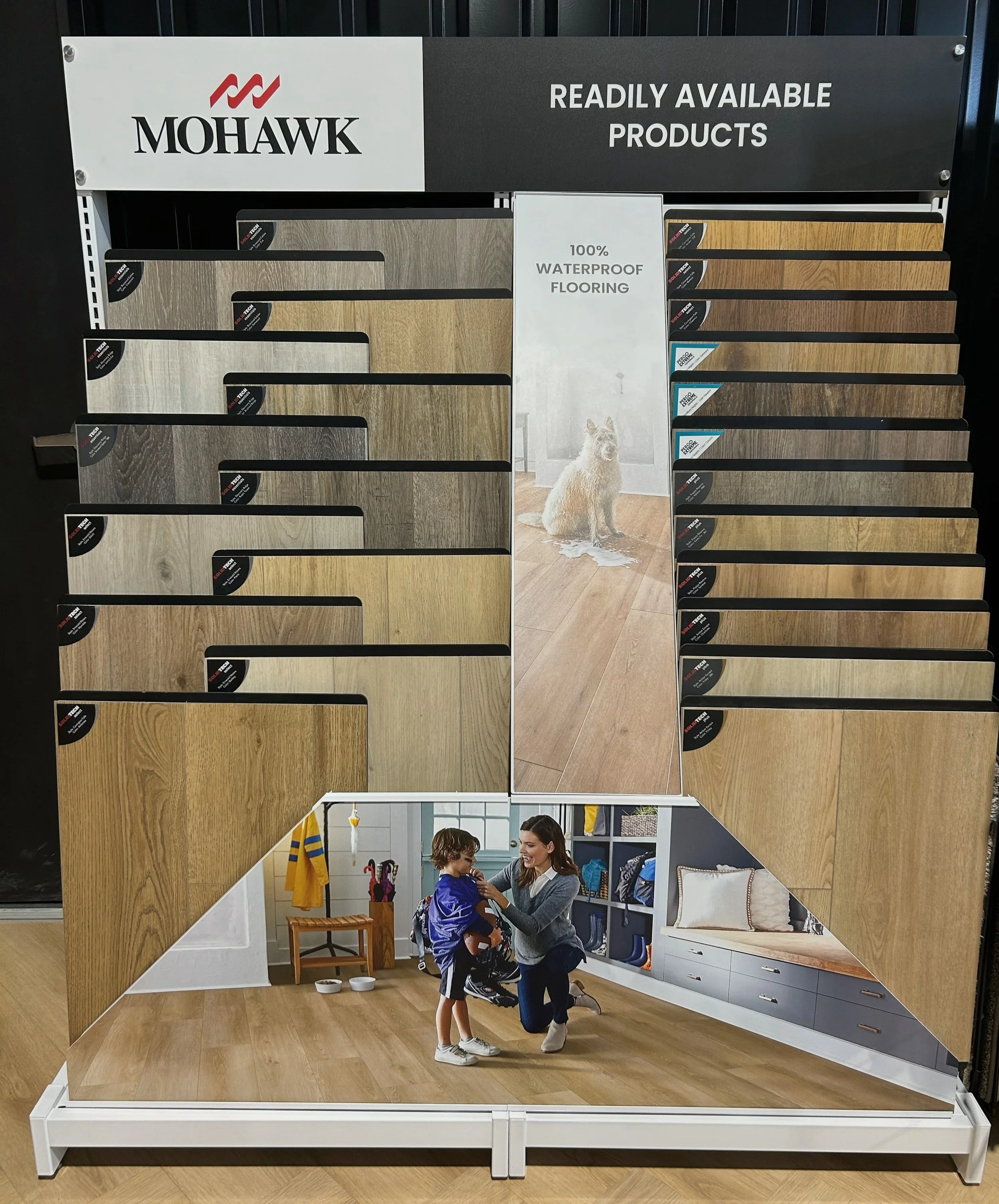 Display of Mohawk wood flooring samples with a picture of a dog on a hardwood floor in the background and a person helping a child try on a backpack in a room exhibit.