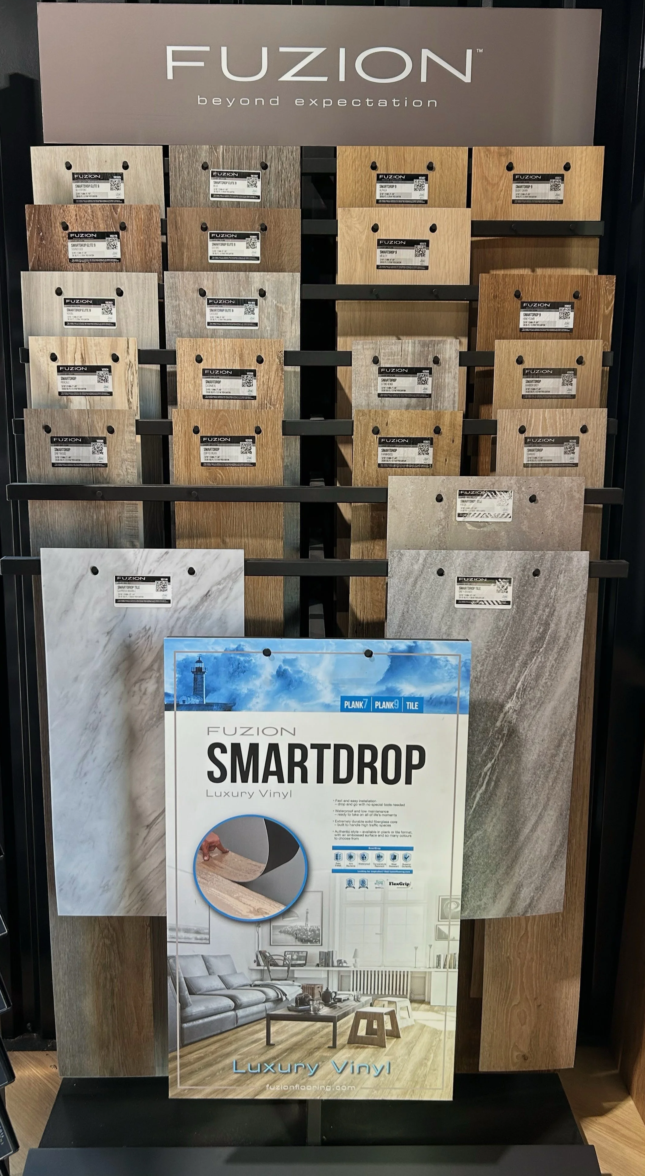 Display of various luxury vinyl plank and tile flooring samples labeled 'Fuzion SMARTDROP'. A large sign above reads 'Fuzion beyond expectation'.