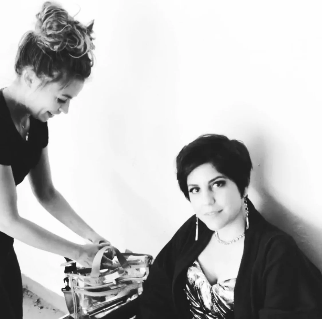 Black and white photo of two women, one with short dark hair and a necklace, sitting and facing the camera, while the other woman with curly hair is smiling and leaning over a bag with various items inside.