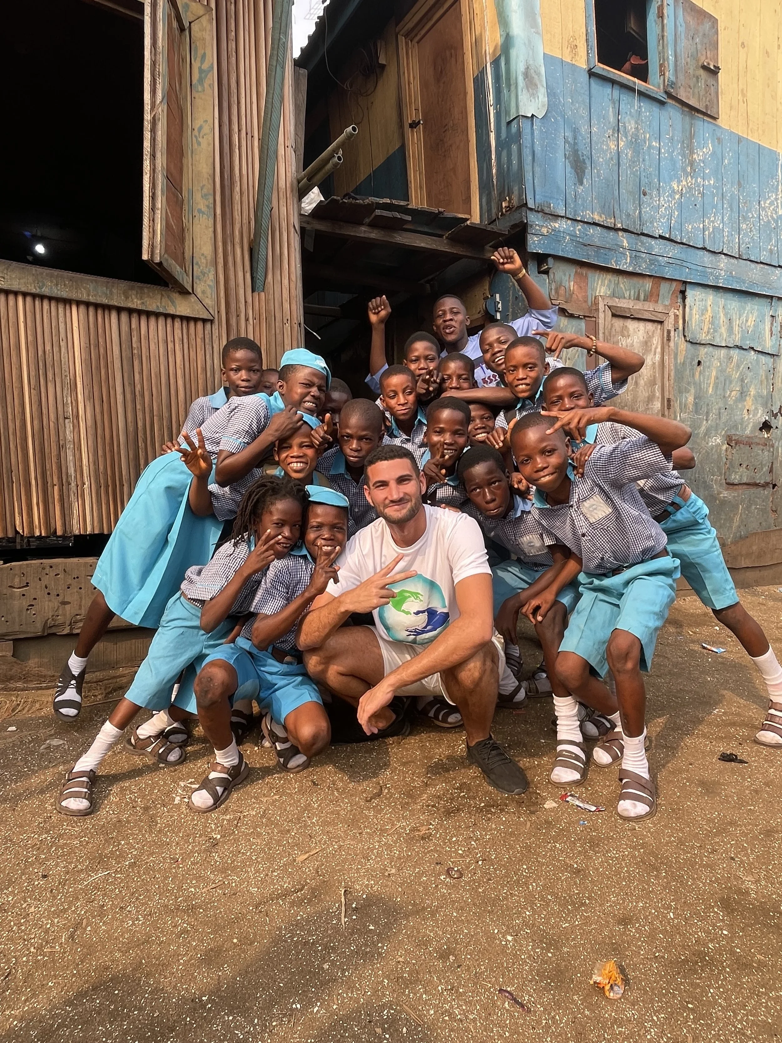 A diverse group of schoolchildren and a man are posing together outdoors in front of a wooden house. The children are wearing school uniforms and making playful gestures, while the man is sitting on the ground and smiling.