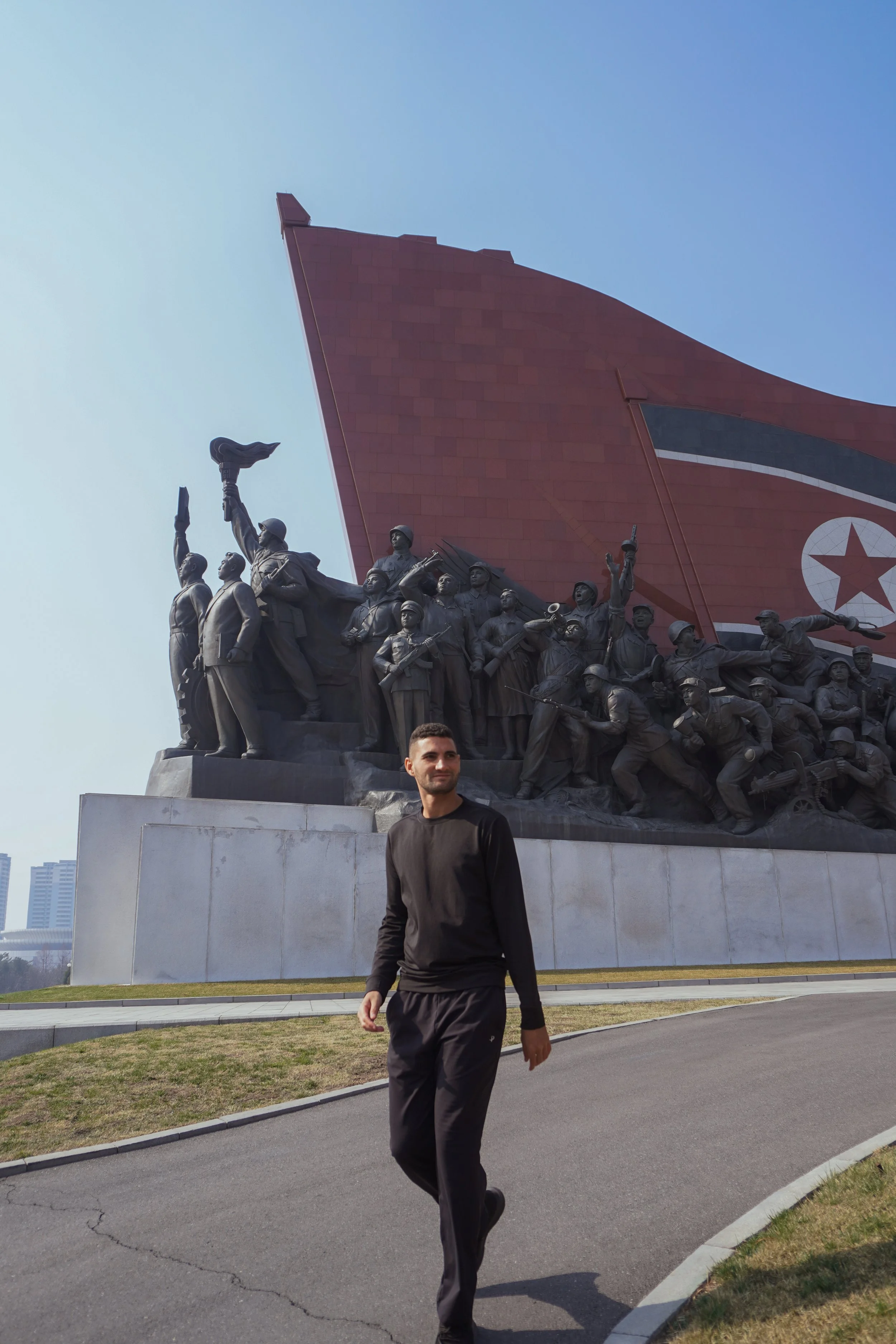 A man walking outdoors in front of a large monument with historical figures and a red flag with a star, under a clear blue sky.
