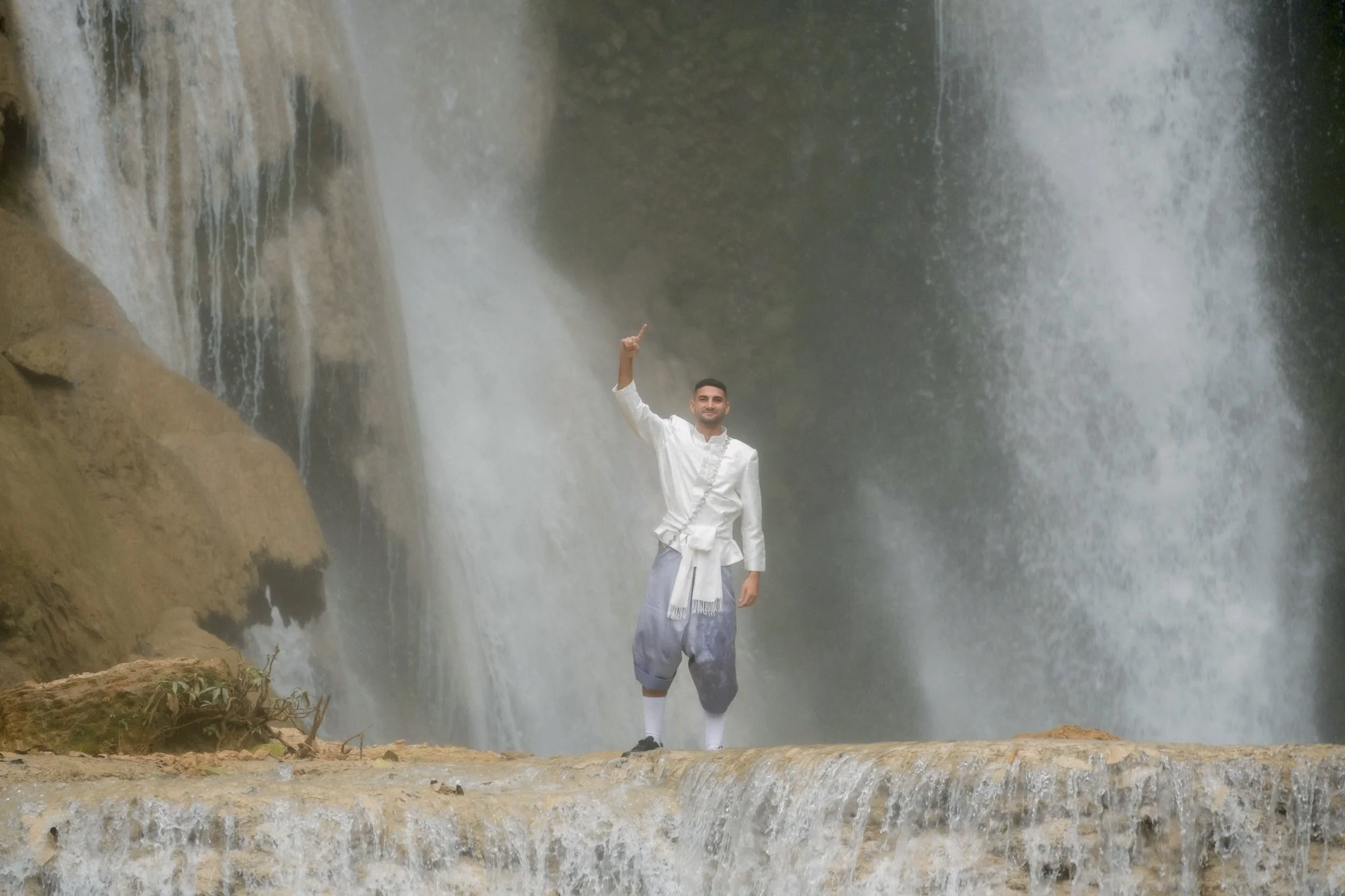 A man in traditional white attire standing on a ledge in front of a waterfall, pointing upward with his right hand.
