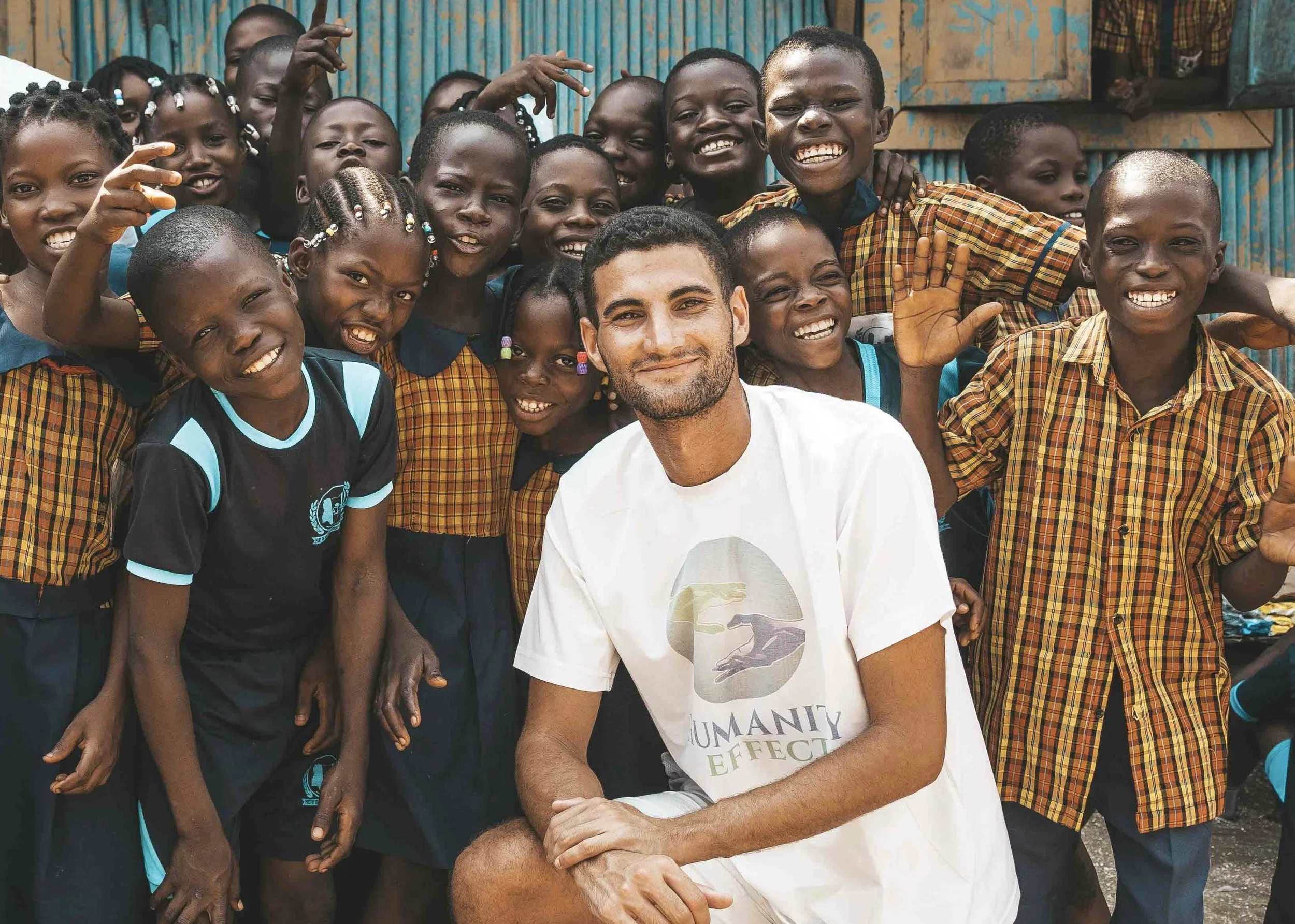 A group of children and a man taking a group selfie outdoors. The children are smiling and some are waving. The background features blue and yellow structures.