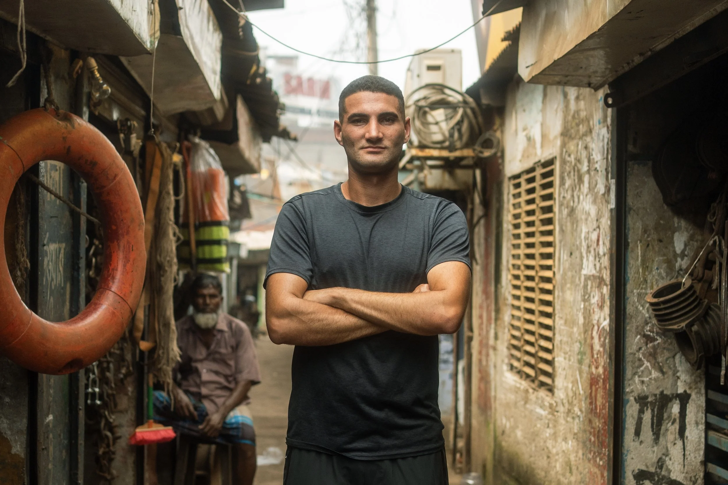 A young man standing with arms crossed in a narrow alley with worn walls, pipes, and hanging wires, with an older man sitting in the background.