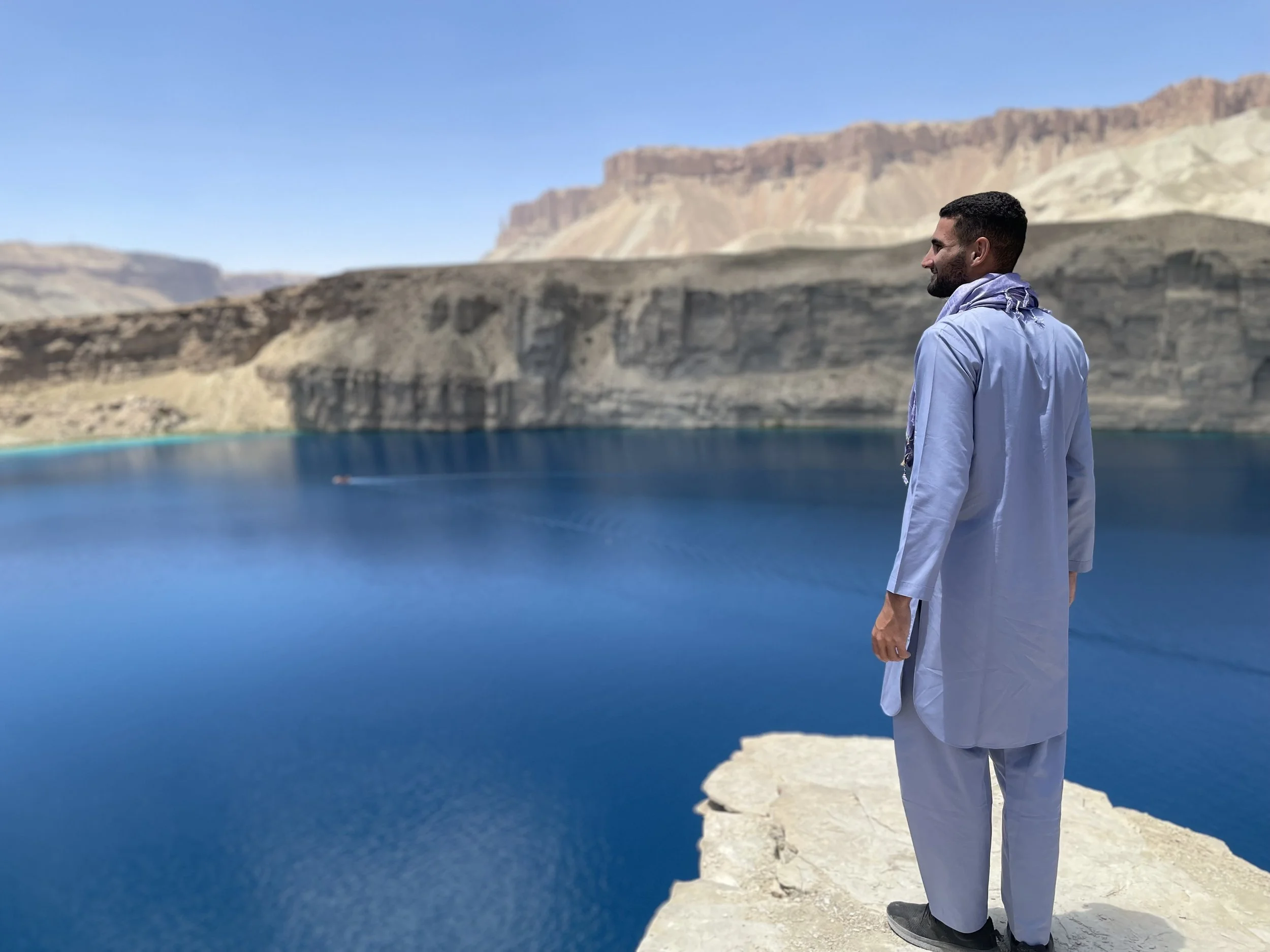 A man dressed in traditional Middle Eastern attire standing on a white rocky outcrop, overlooking a deep blue body of water with rugged, arid mountains in the background under a clear blue sky.
