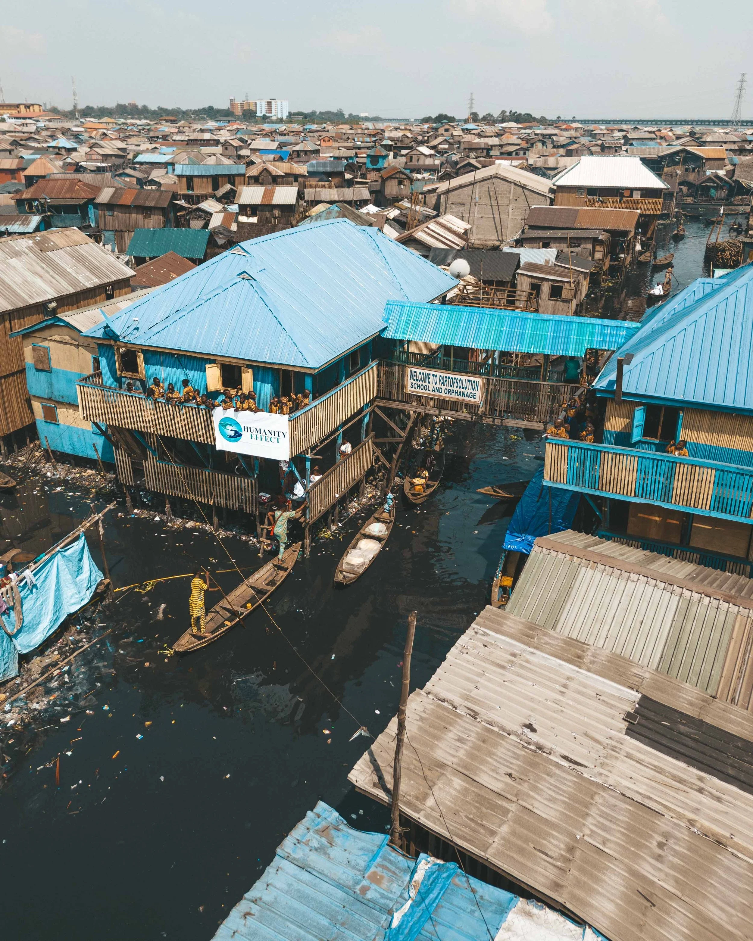 Aerial view of a densely populated slum with wooden and metal houses built on stilts over a waterway, some with blue tin roofs, and boats floating alongside. A sign reads "Welcome to Parto Solution School and Orphanage."