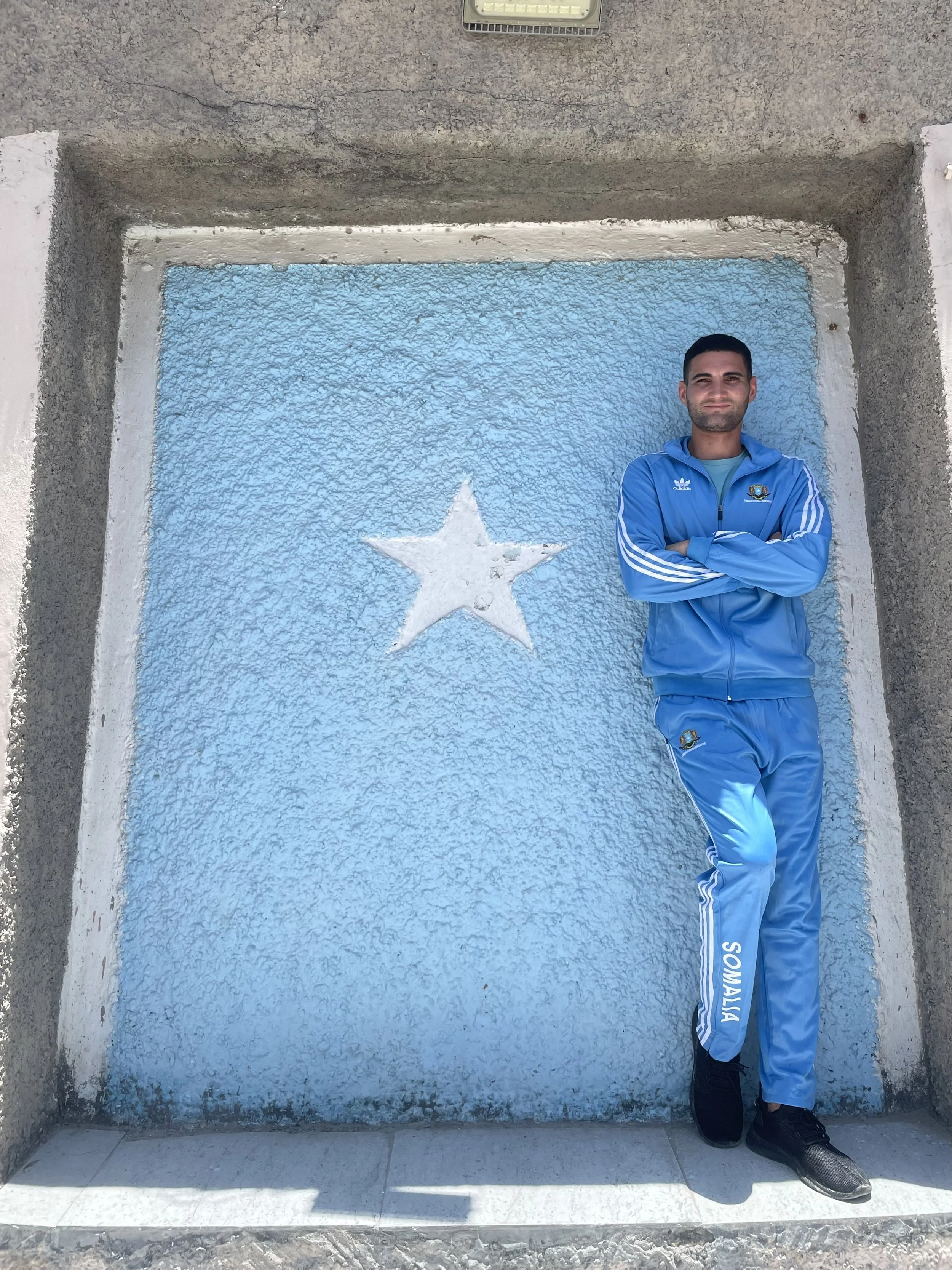 A man in a blue tracksuit with the word 'Sonalia' on the leg, standing with arms crossed, smiling, in front of a blue wall with a white star painted on it. The wall has a white border and a grey concrete surround. The man is wearing black athletic shoes.