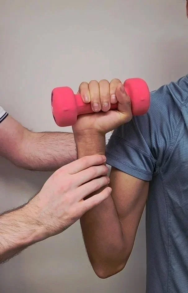 Person in a blue shirt having their arm measured with a pink dumbbell, with an individual helping to hold the arm steady.