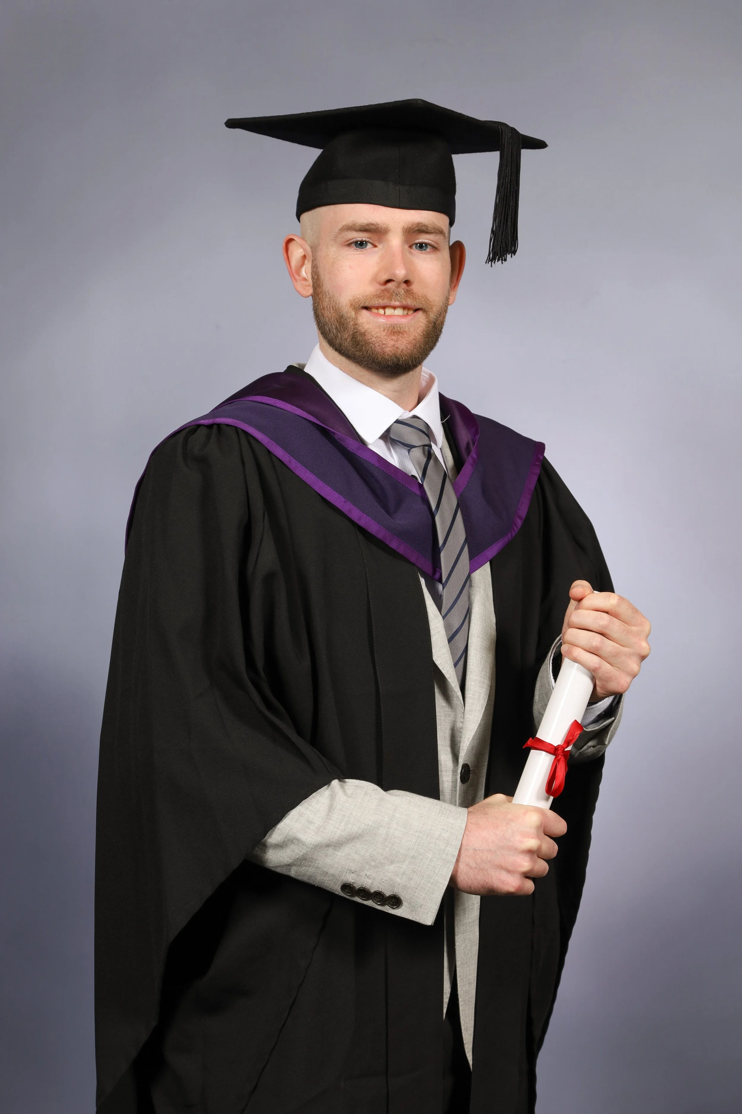 A young man in graduation attire, including a black cap and gown with a purple and black hood, holding a diploma tied with a red ribbon, smiling at the camera.