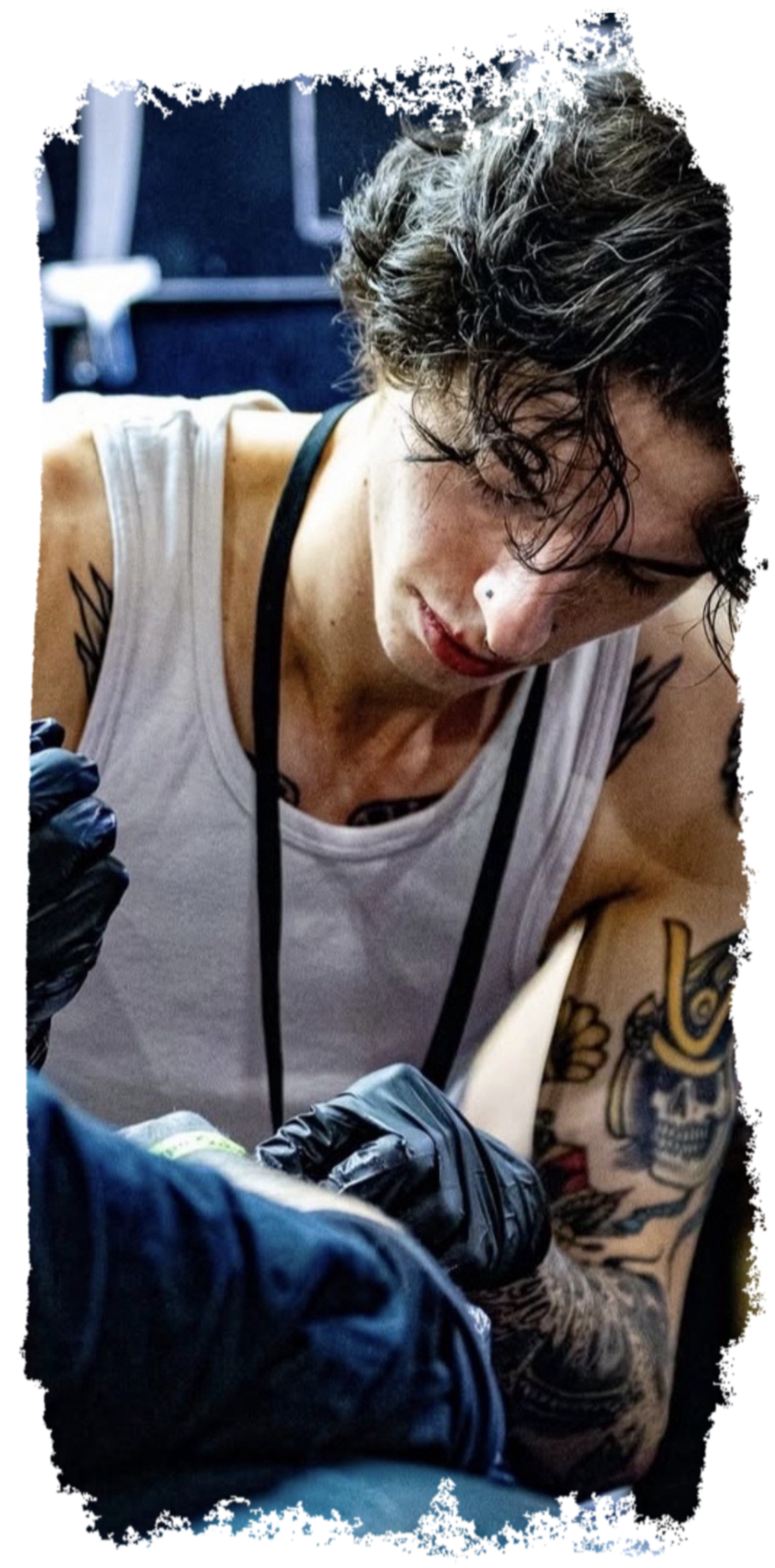 A woman with tattoos on her arms, black curly hair, and a nose piercing is seen tattooing someone's arm. She is wearing a white tank top and black gloves.