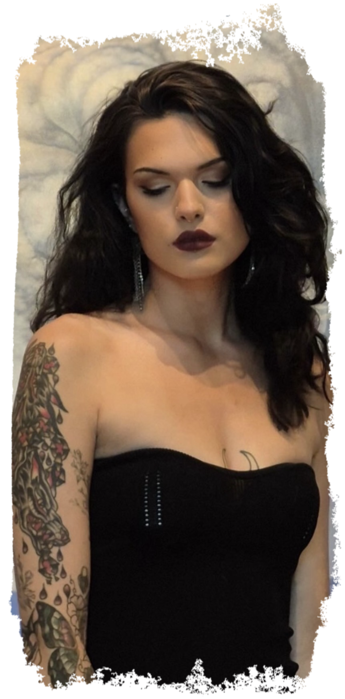 A woman with dark, curly hair and tattoos on her left arm, wearing a black strapless top, looking down with closed eyes.