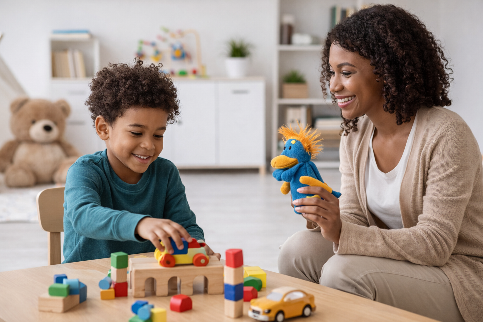 A young boy and a woman, likely his mother or teacher, playing with colorful toys at a table in a bright, organized playroom. The boy is stacking wooden blocks and a toy car, while the woman holds a blue plush puppet with orange hair, smiling at the boy. In the background, there is a large teddy bear and shelves with books and plants.