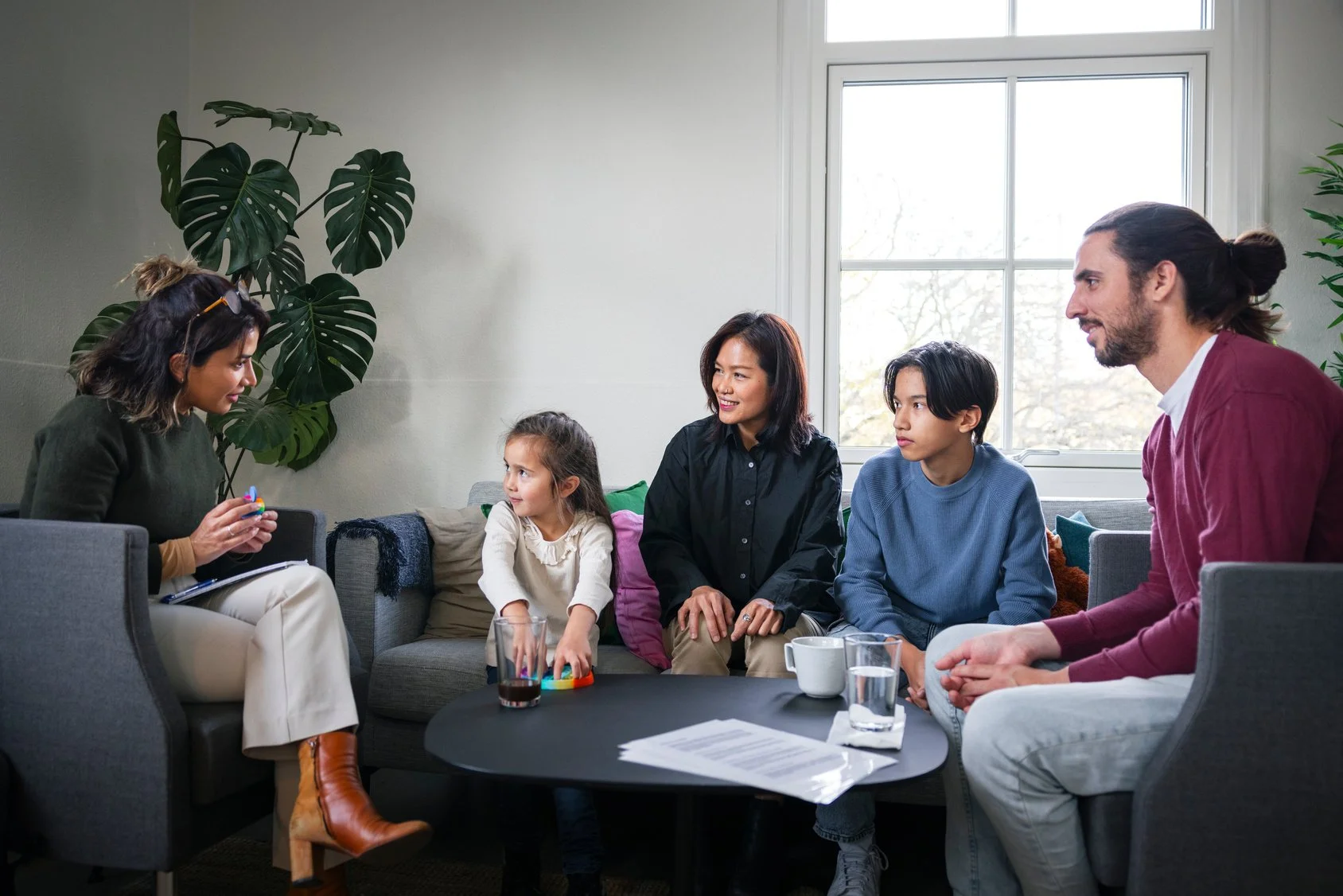 A group therapy session with five people sitting on a sofa and talking, including a woman with a notepad, a young girl, a woman with black hair, a teenage boy, and a man with long hair and beard, in a well-lit living room with a large window.