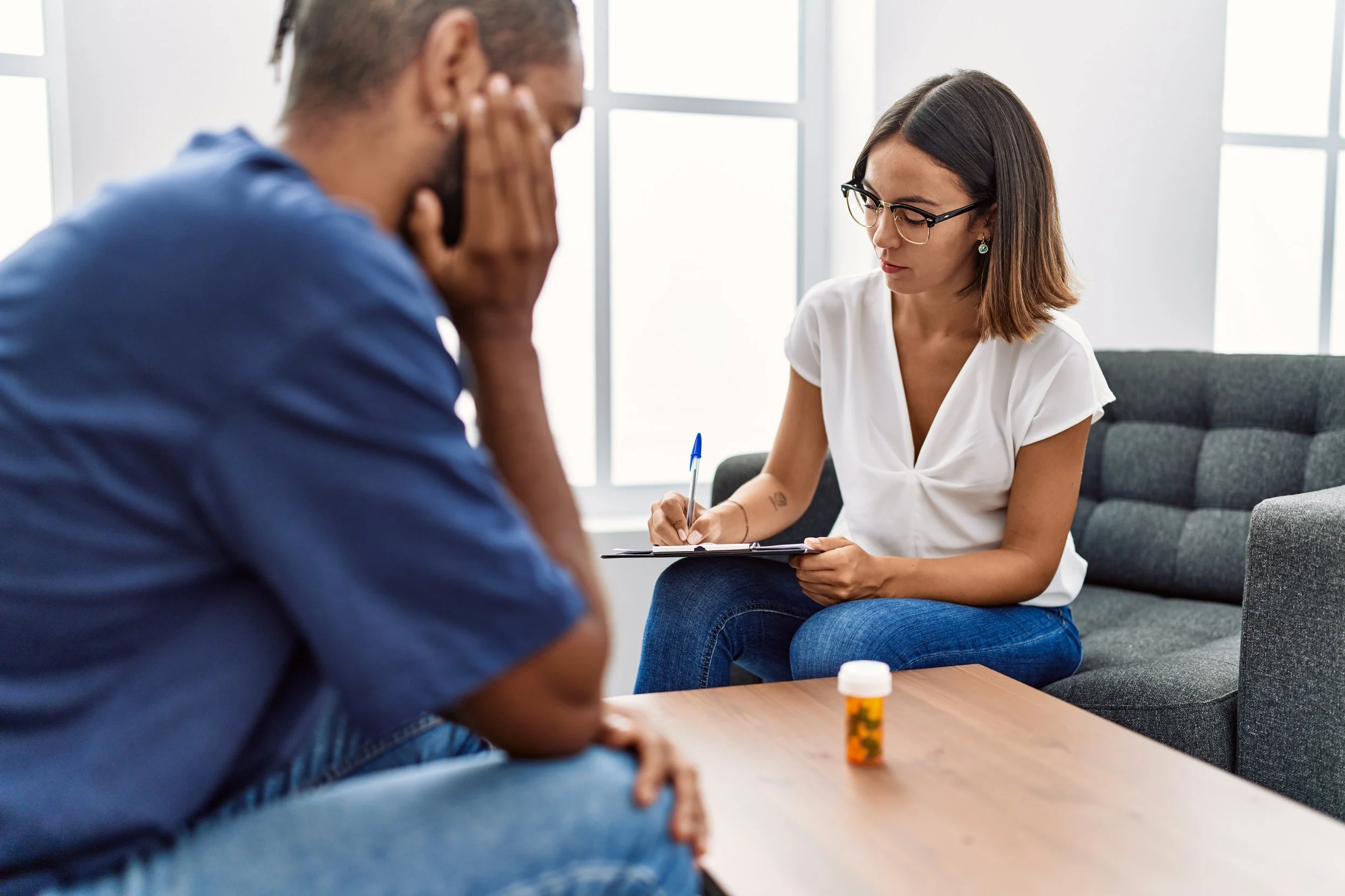 A woman in glasses taking notes during a therapy session with a man sitting across from her, distressed, in a bright office with large windows and a bottle of medication on the table.