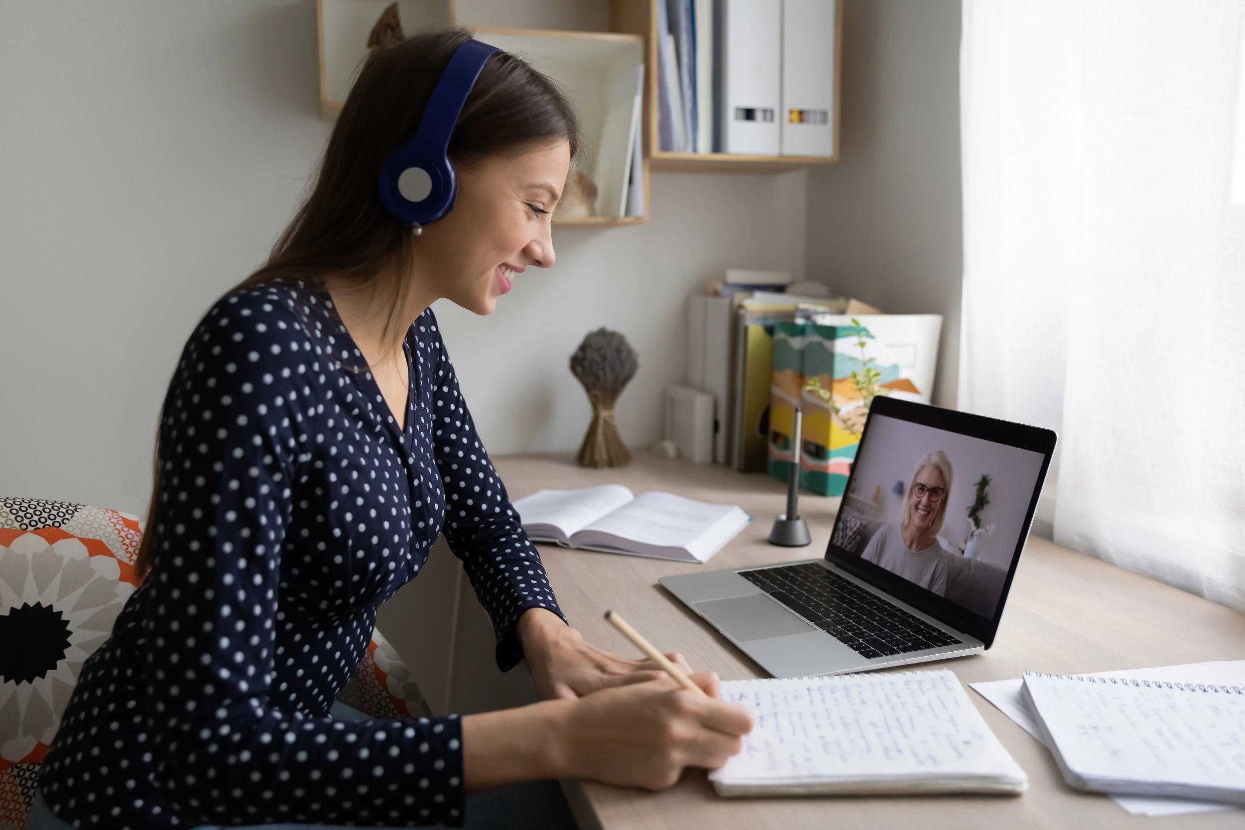 Woman with brown hair wearing a navy blue polka dot shirt, listening to music with headphones, taking notes at a desk while on a video call with a woman on a laptop.