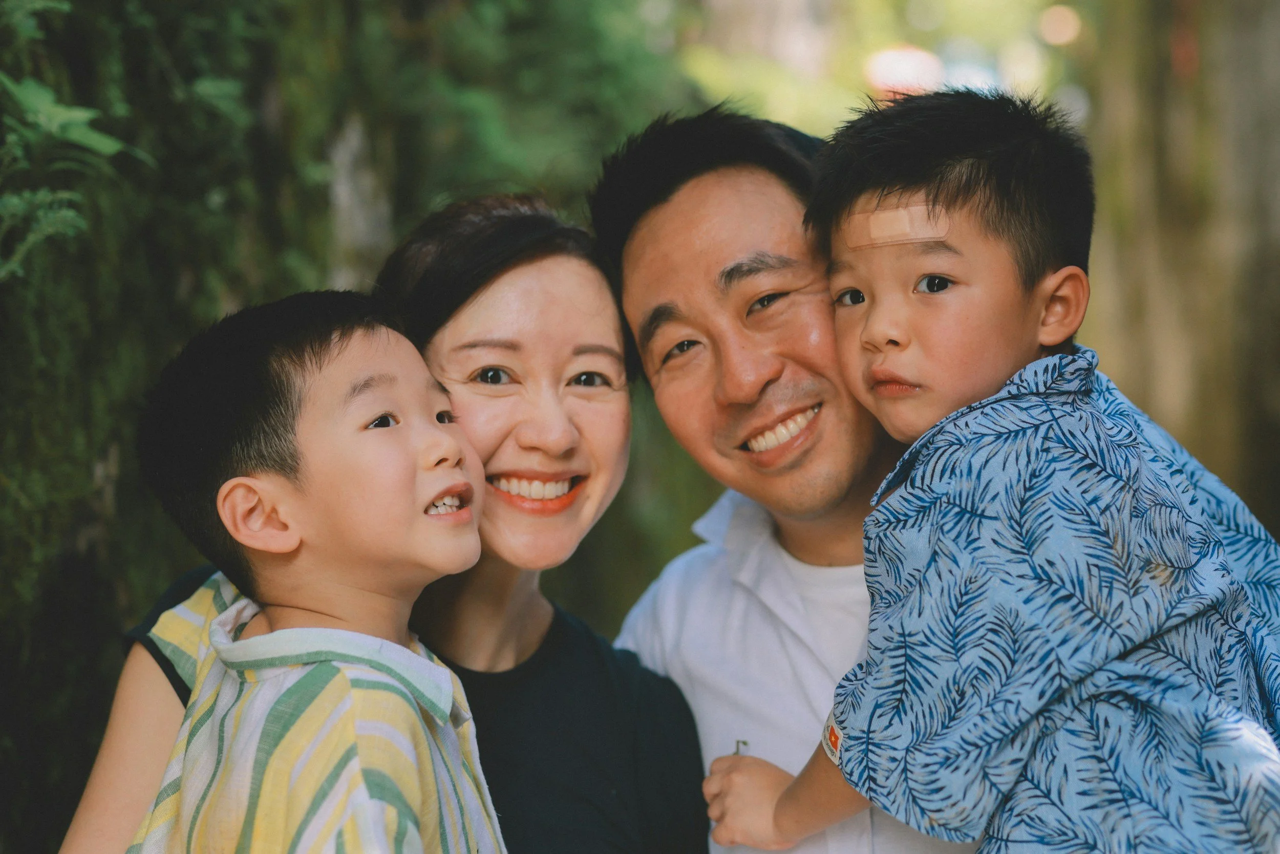 A happy family of four smiling and hugging outdoors in a green, natural setting.