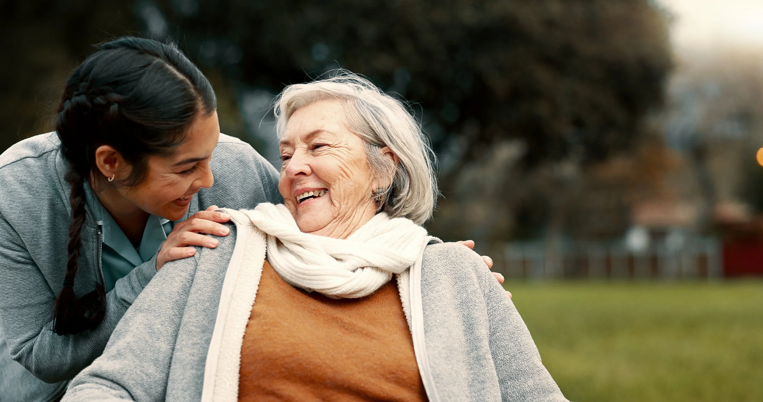 A woman with dark hair and a braid, wearing a gray jacket and school uniform, hugs an elderly woman with gray hair, wearing a gray hoodie, orange shirt, and white scarf, outdoors in a park, both smiling and enjoying each other's company.