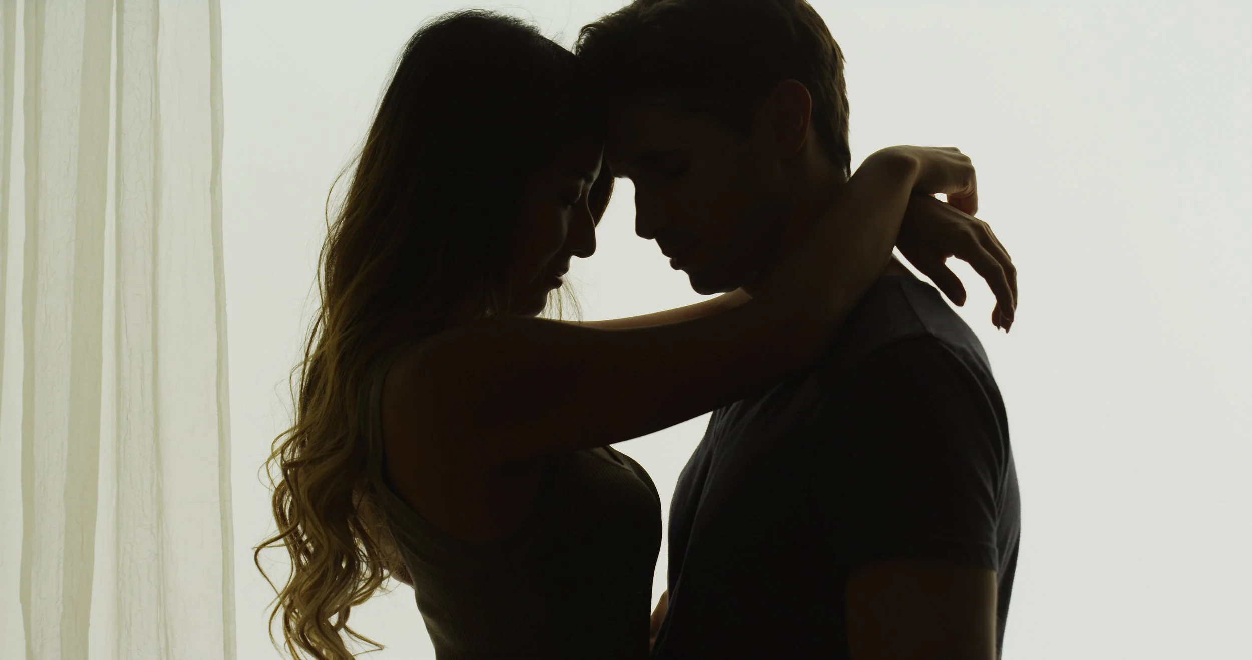Silhouette of a man and a woman with foreheads touching and arms around each other in front of a backlit curtain.