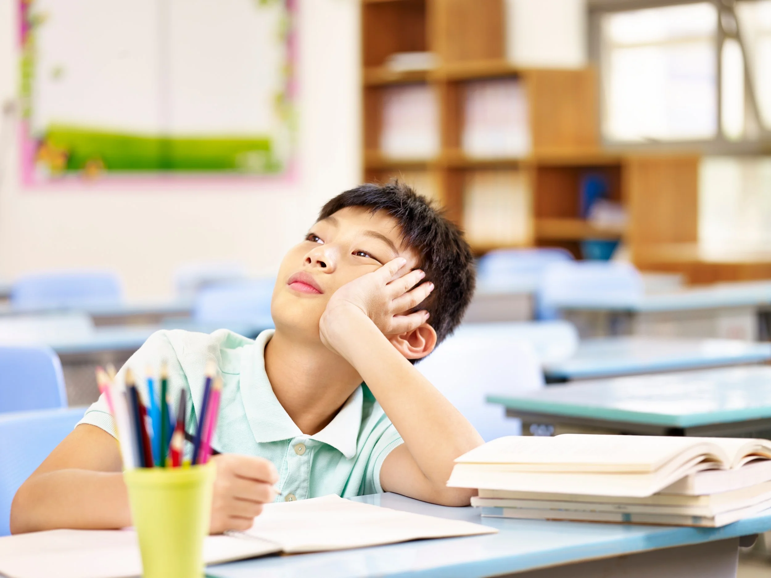 A boy sitting at a classroom desk with an open book and a yellow container holding colored pencils, looking bored or distracted.