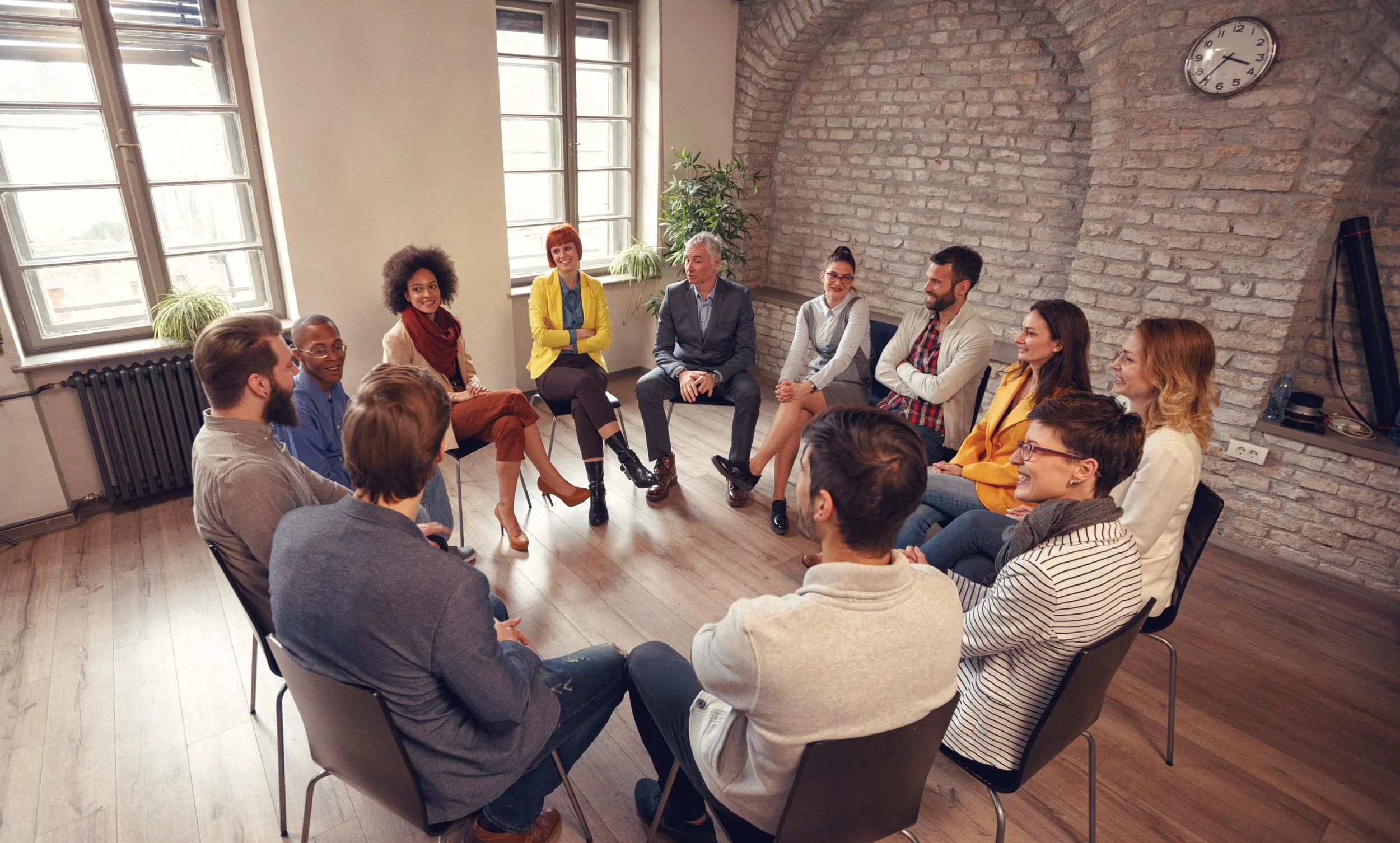 A diverse group of people sitting in a circle during a meeting or discussion in a bright, brick-walled room with large windows.