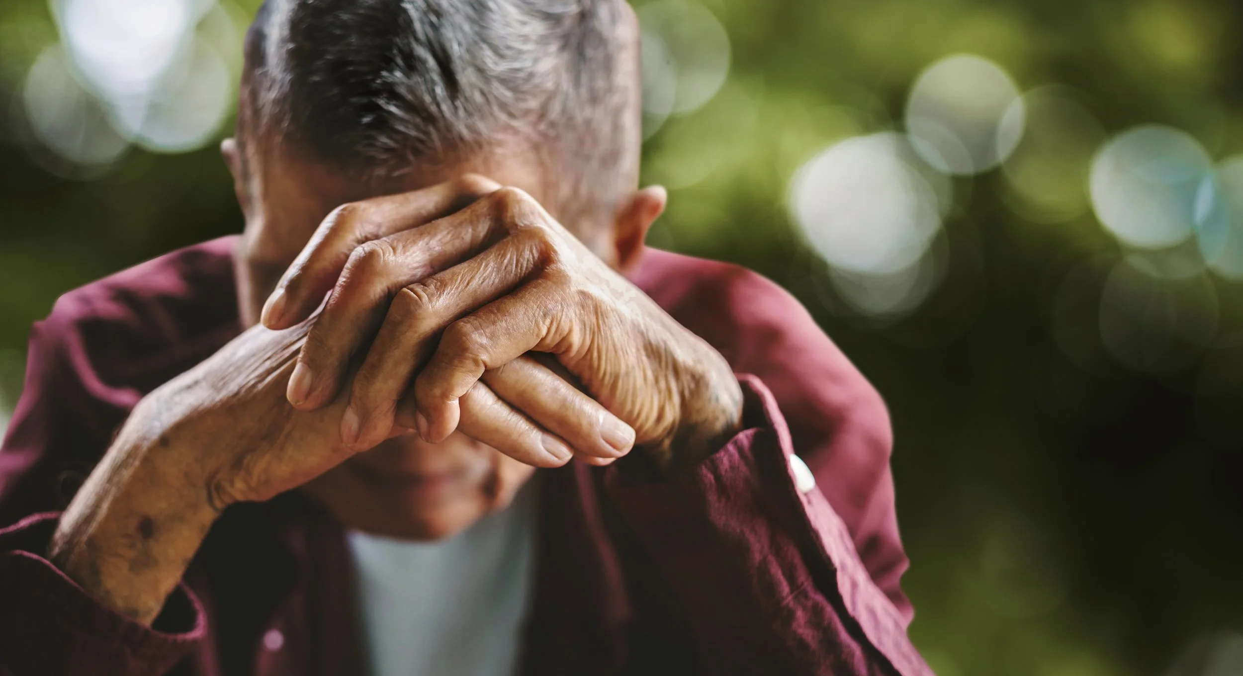 An elderly person with gray hair is sitting outside with hands clasped over face, appearing to be in deep thought or sadness, with a blurred green foliage background.