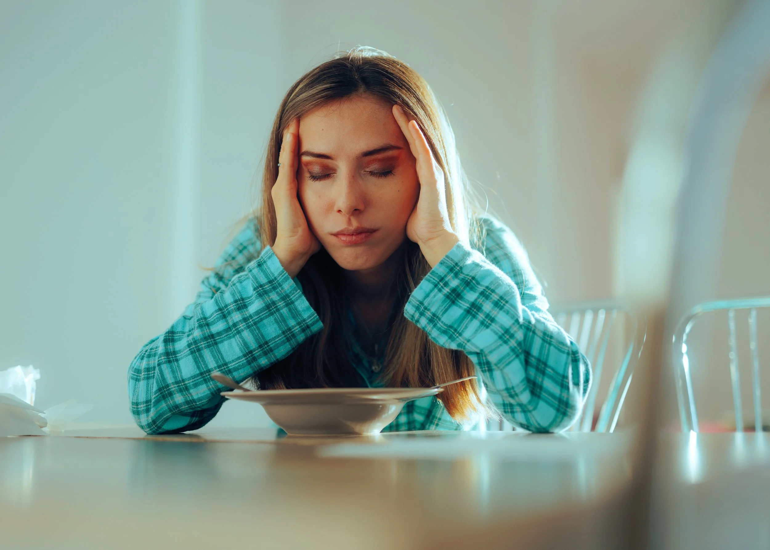 Young woman in plaid shirt sitting at table with hands on her head and a sad expression, with an empty bowl in front of her.