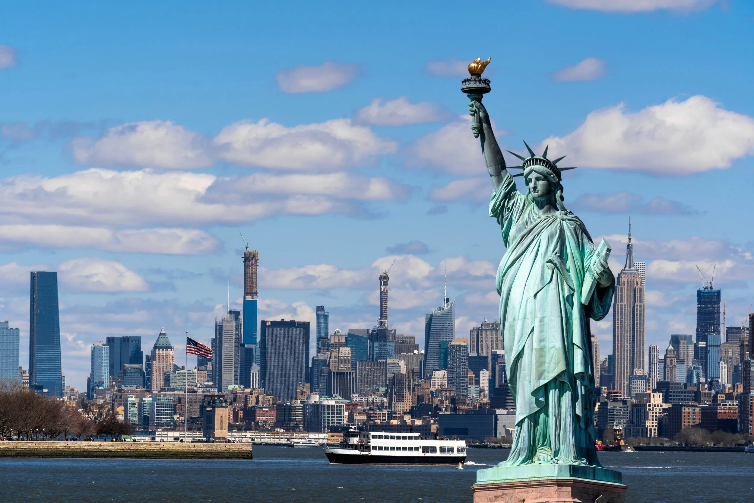 The Statue of Liberty overlooking the New York City skyline with skyscrapers, a boat on the water, and a partly cloudy sky.