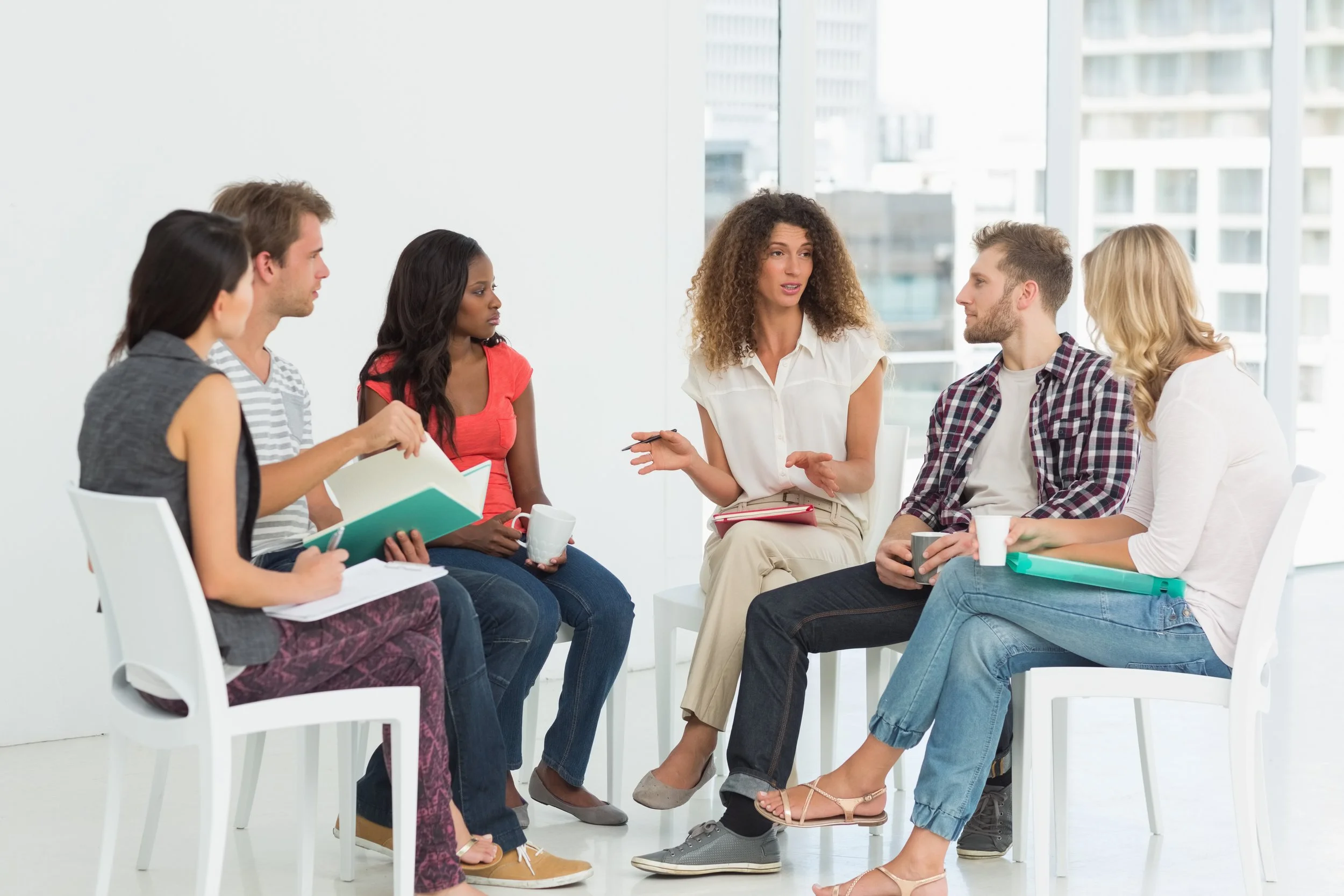 A diverse group of six people sitting in a circle having a meeting or discussion in a bright office space with large windows.