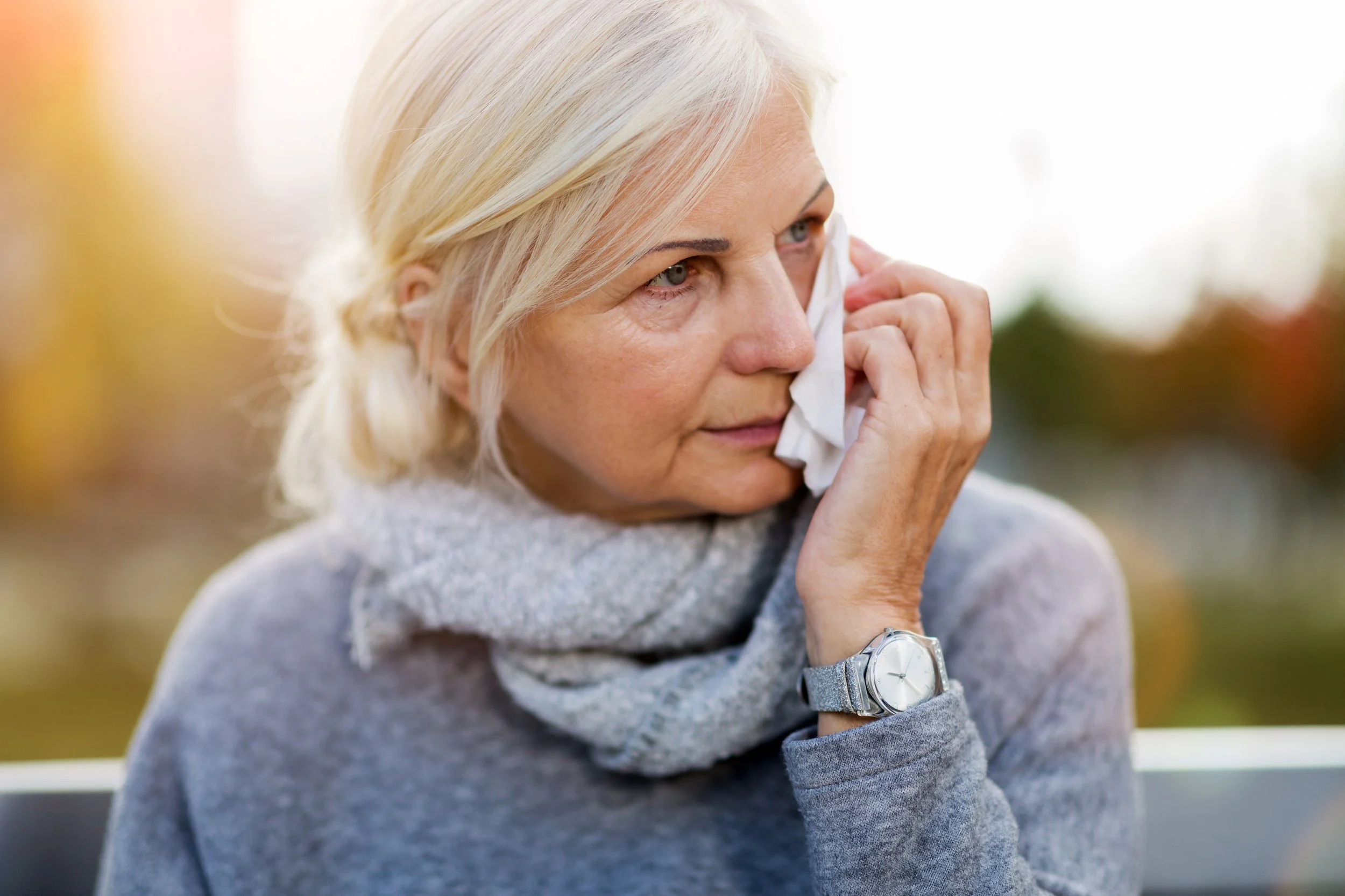 A woman with blonde hair in a ponytail wiping tears from her face with a tissue outdoors in autumn weather.