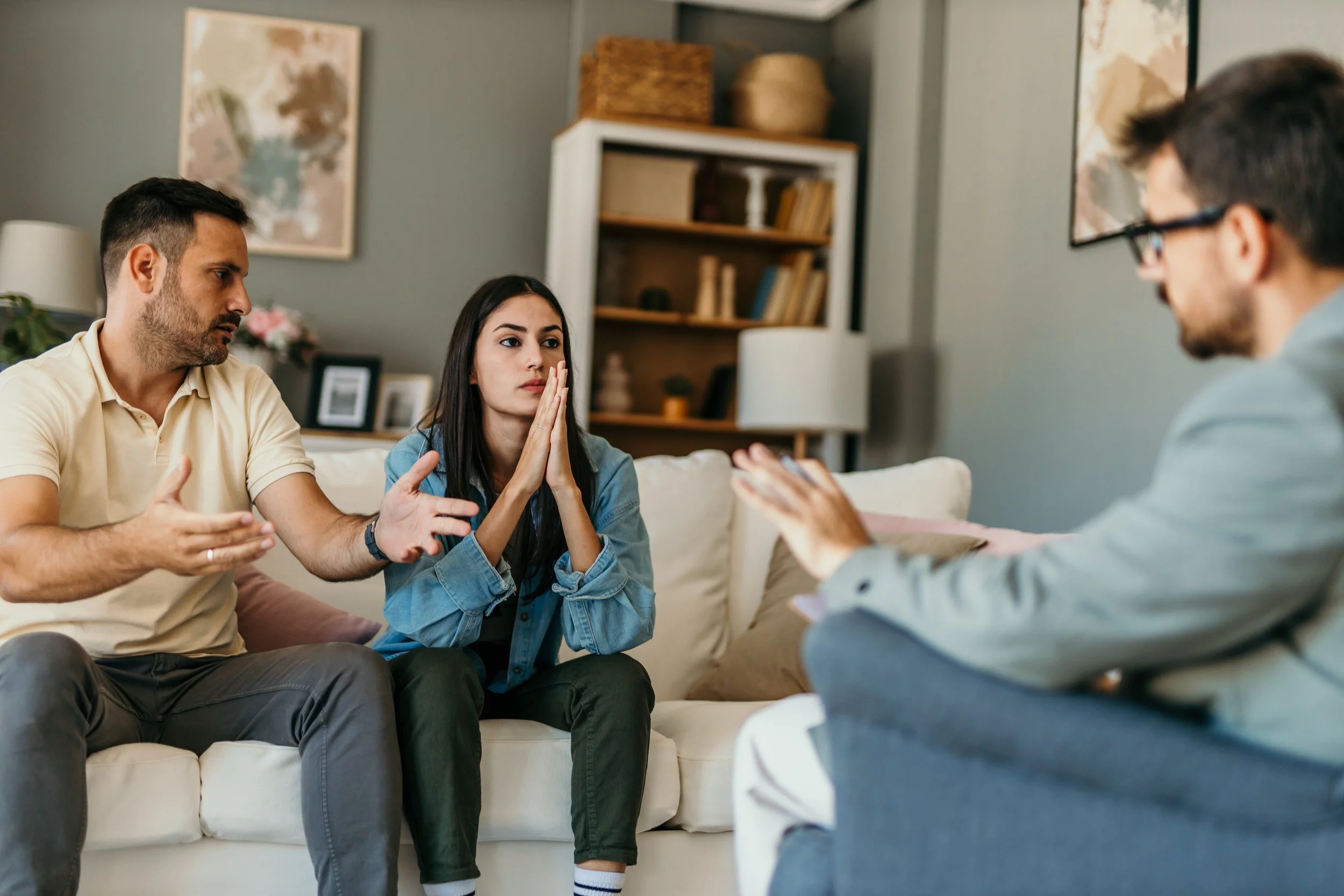 A young woman with long dark hair sits on a sofa with her hands pressed together as she listens to a man talking. Next to her, a man with short dark hair gestures with his hands. Across from them, a man with glasses and dark hair talks and uses hand gestures. The scene appears like a counseling or therapy session in a cozy living room with framed art and books around.
