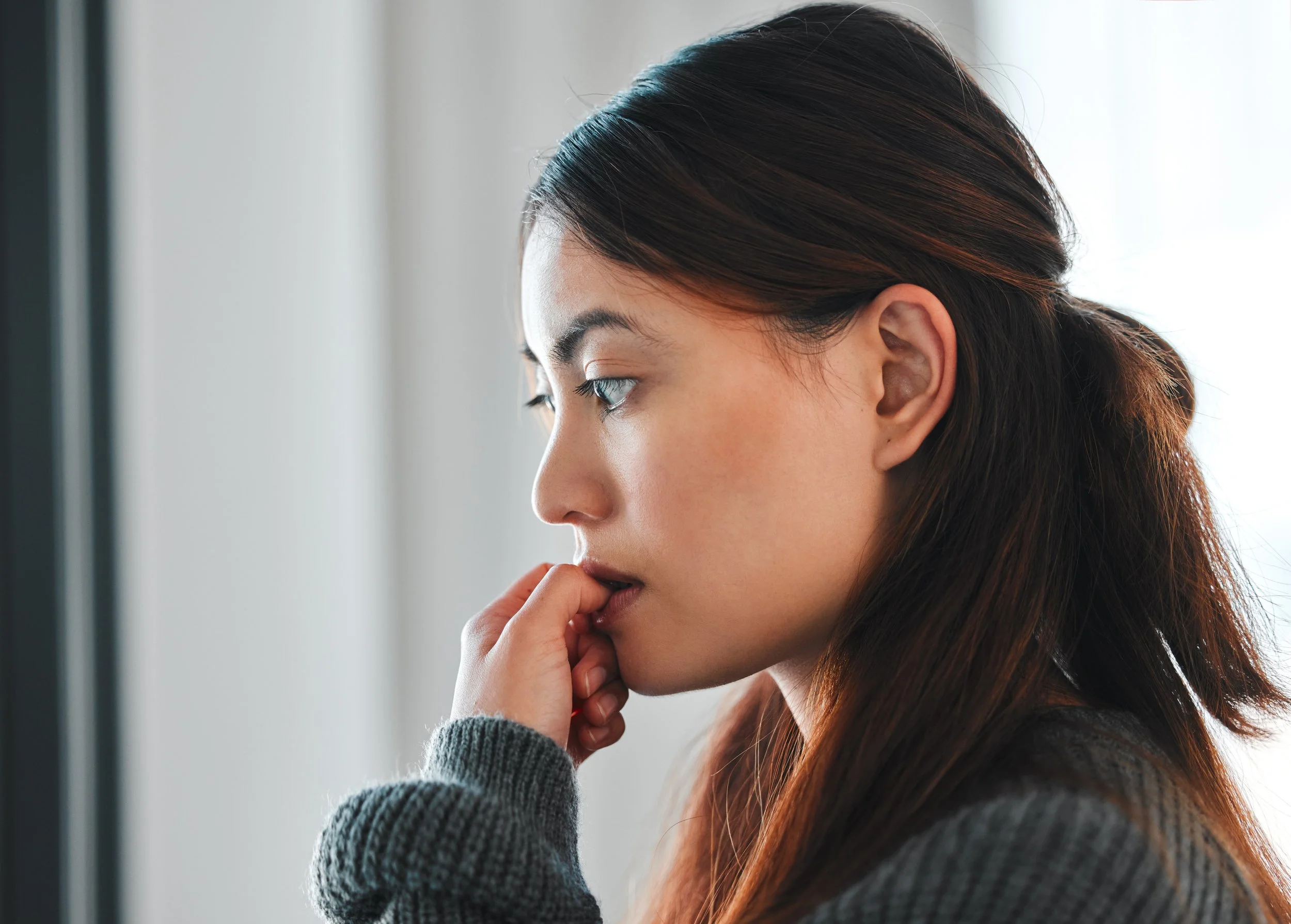 A young woman with long dark hair looking out a window, with her hand near her mouth, wearing a gray sweater.