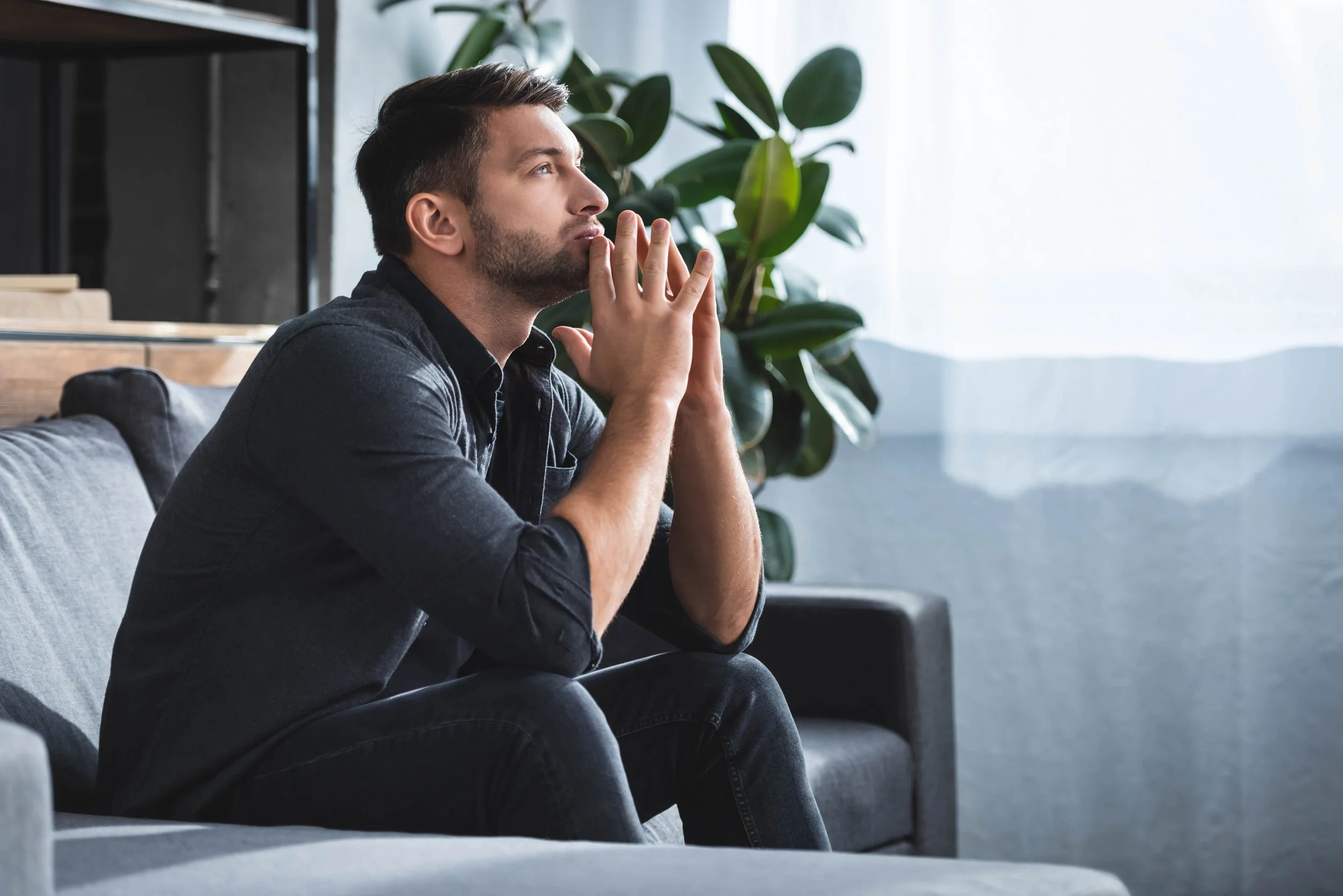 A man sitting on a gray couch with his hands clasped together near his face, looking contemplative, in a bright room with a large plant and white window curtain in the background.