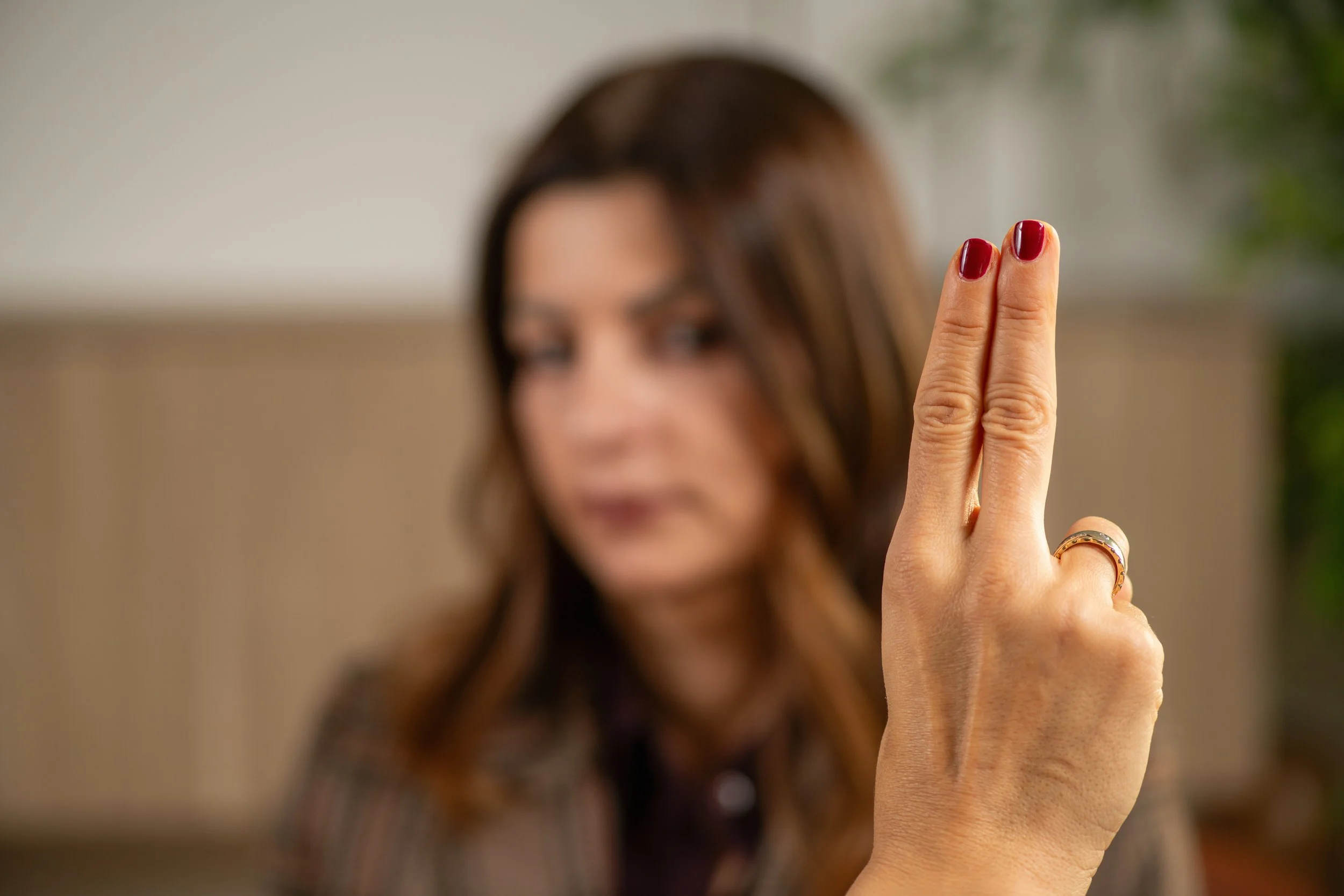 A woman with brown hair, blurred in the background, holds up her hand with two fingers extended in a peace sign or gesture in focus, showing her red-painted fingernails and rings on her fingers.