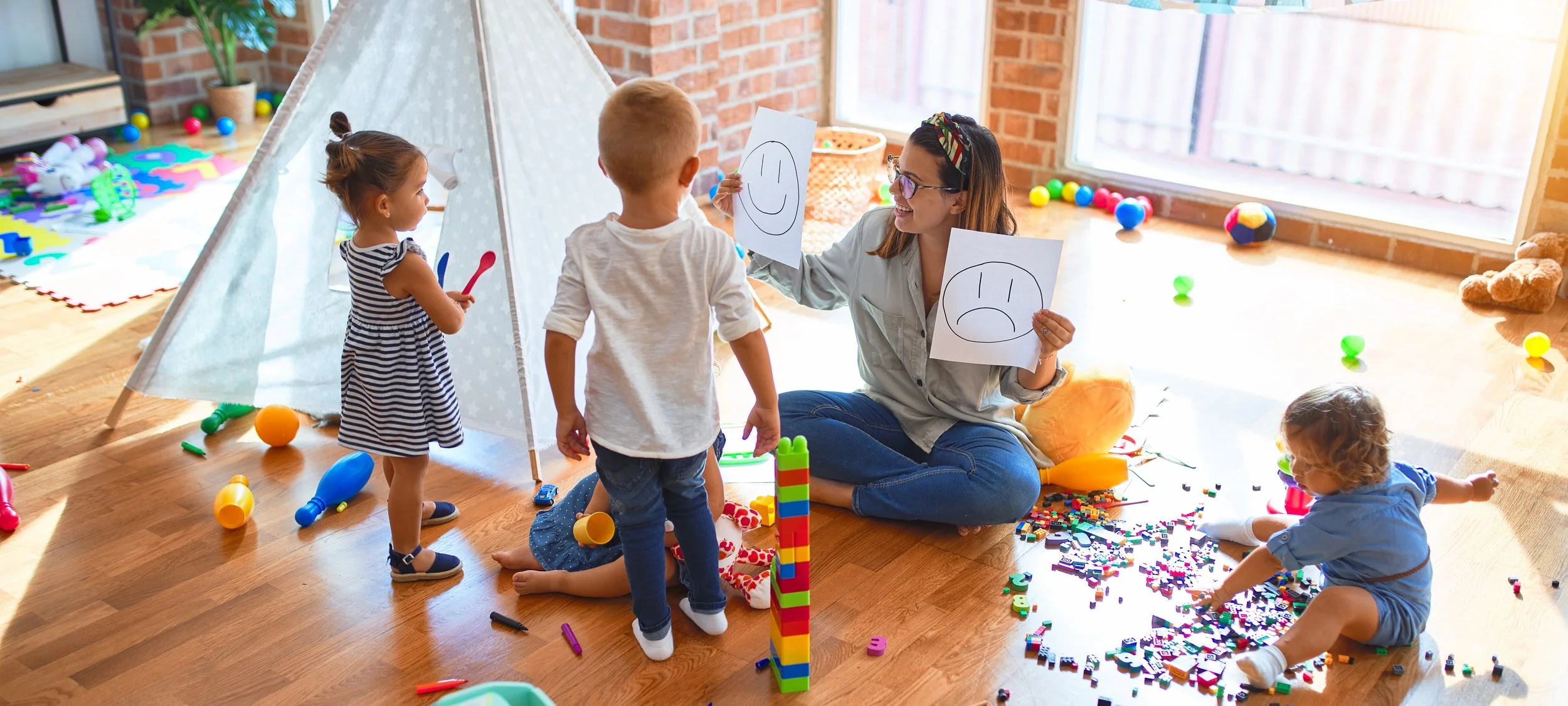 Children and a woman playing with toys in a brightly lit indoor space, with a small tent, scattered toys, and colorful objects on the floor after playing.