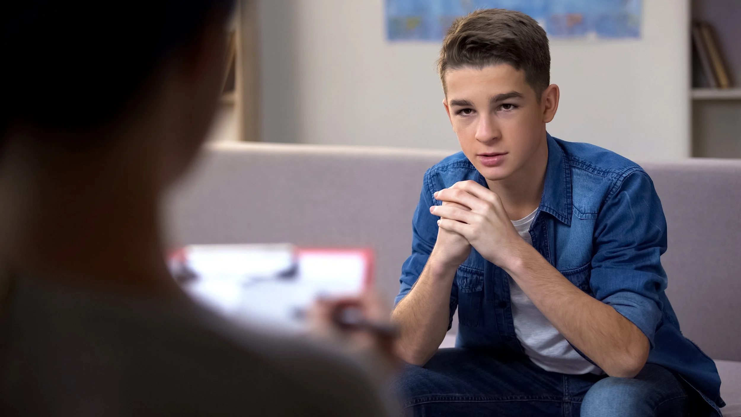A teenage boy with short brown hair, wearing a denim jacket and white shirt, sitting on a gray couch with hands clasped, talking to a woman who is out of focus in the foreground, in a living room with bookshelves and a world map on the wall.