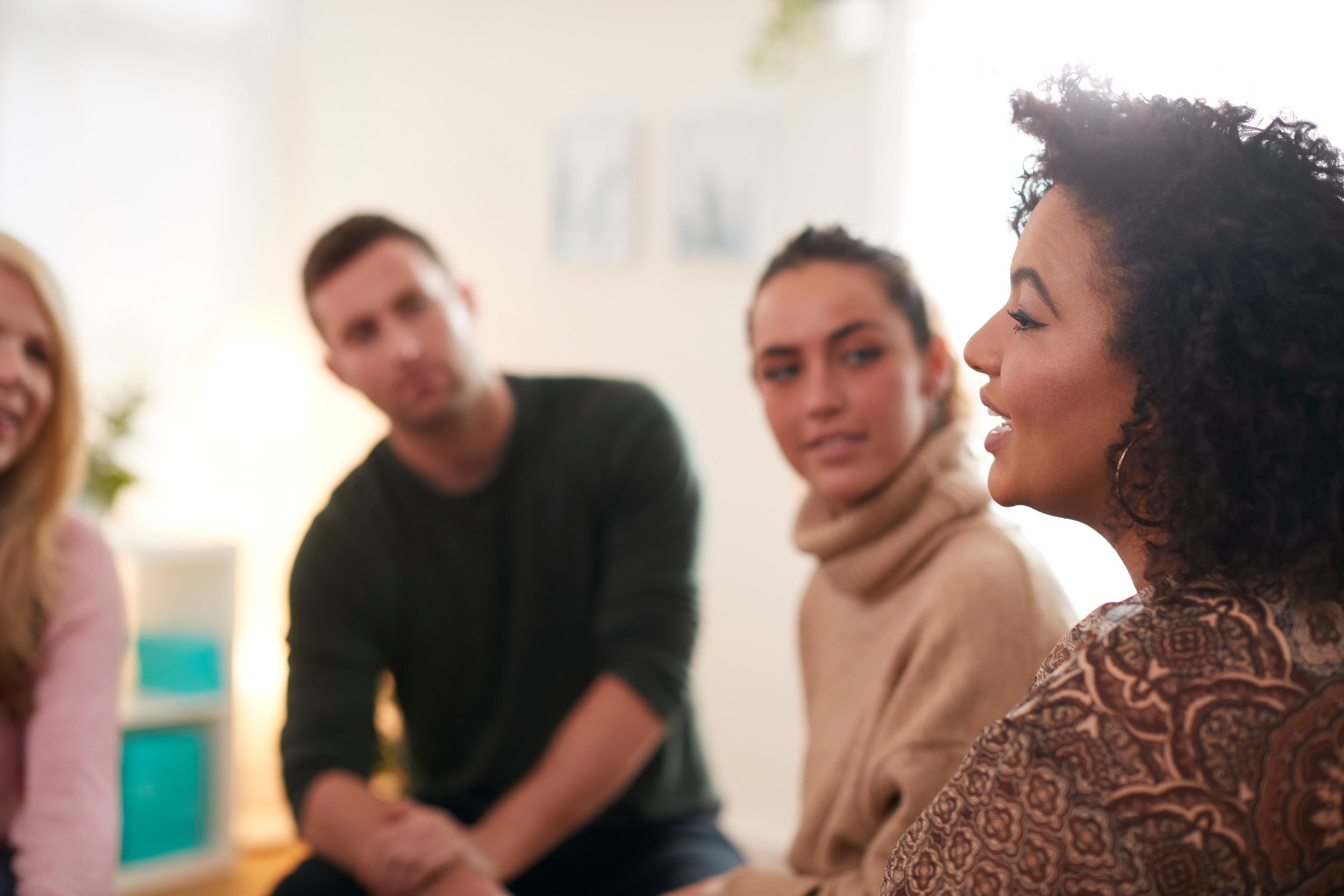 A woman with curly hair speaking or listening in a group therapy session, with three people listening attentively to her, in a bright room.