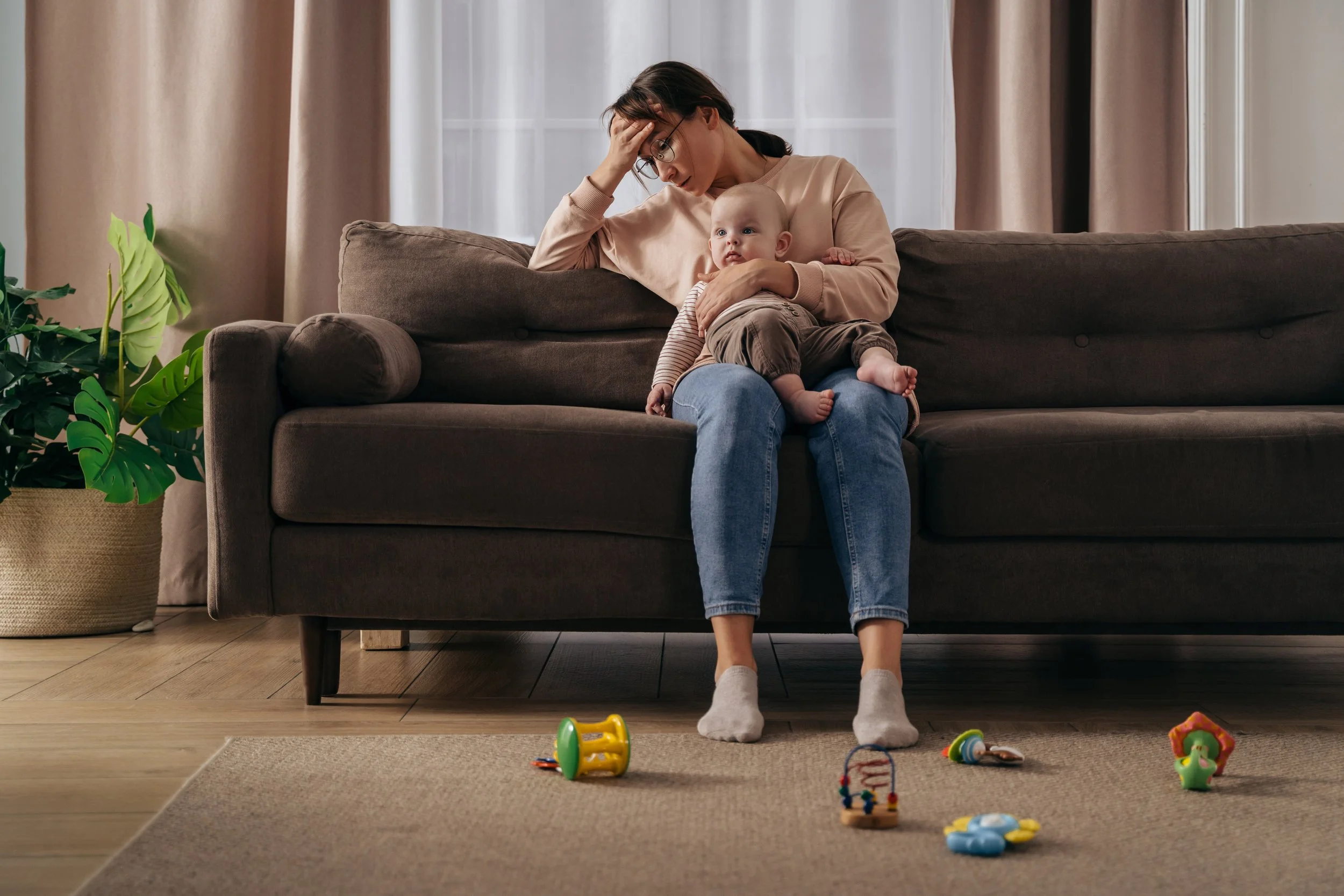 A woman sitting on a dark gray sofa holding a baby on her lap, with her hand on her forehead, looking distressed. The baby is looking to the side. There are colorful baby toys on the beige rug in front of them and a potted plant to the left. The background shows a window with sheer curtains and beige curtains on either side.
