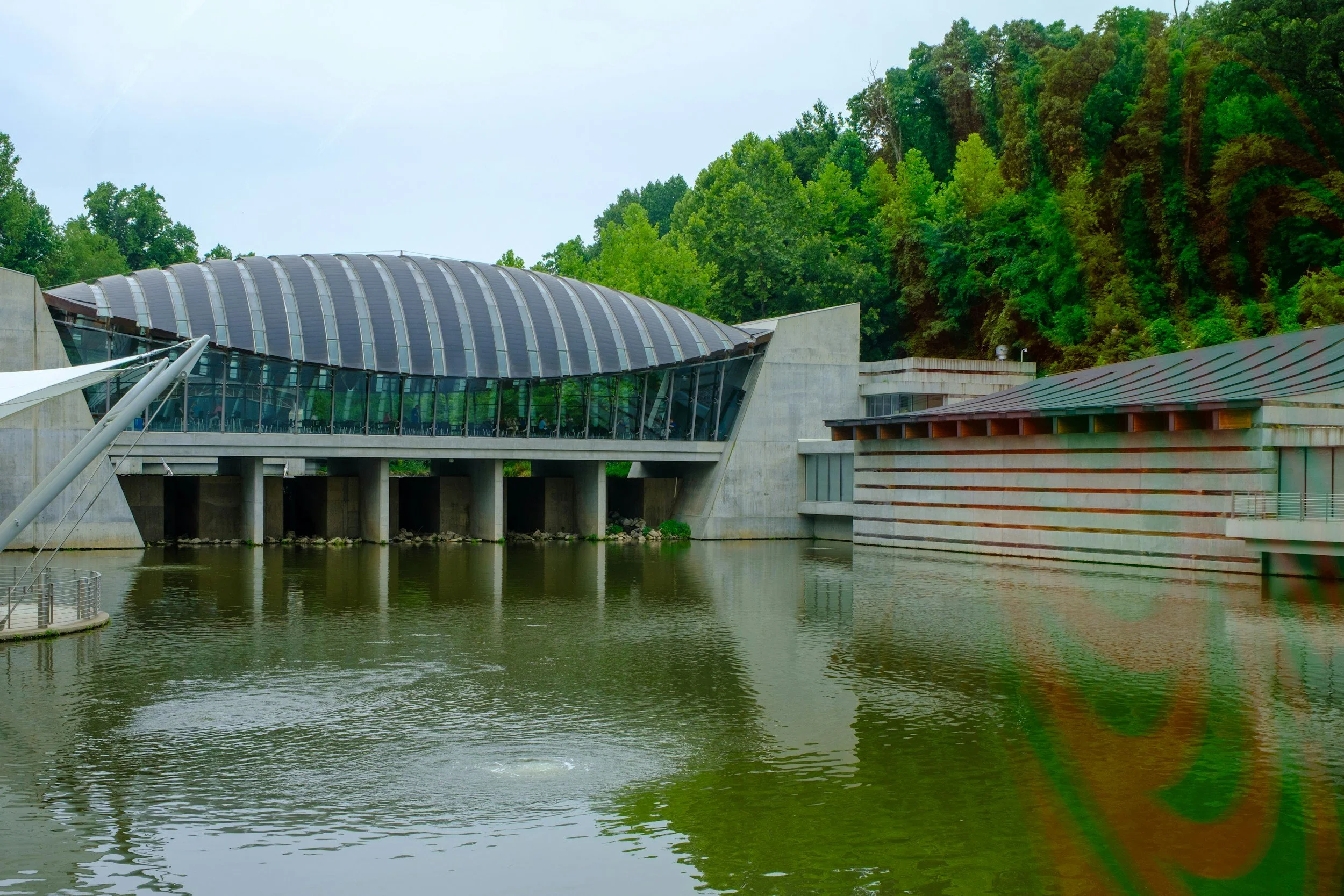 Modern building with a curved roof made of glass panels, situated next to a body of water with green, forested hills in the background.