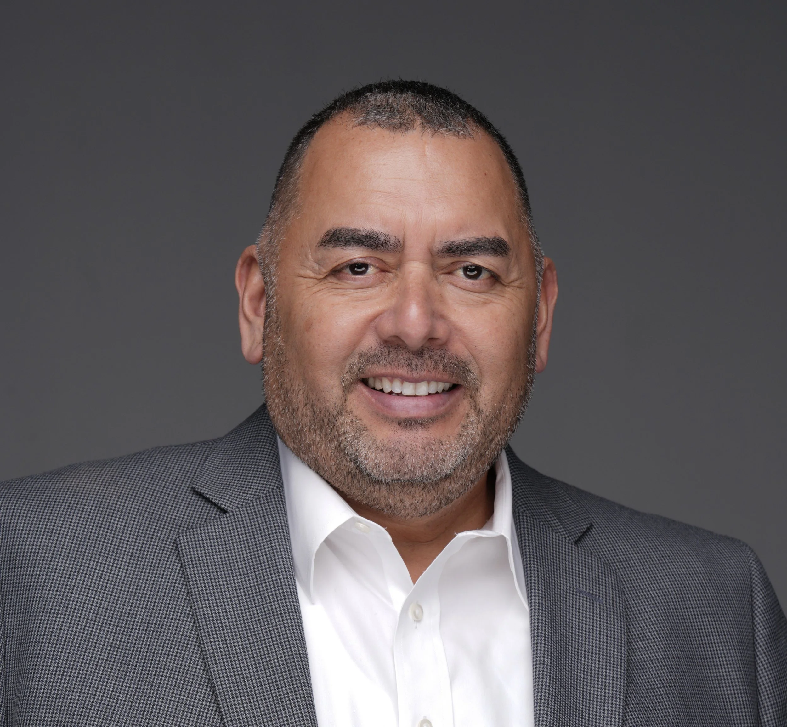 Head and shoulders of a middle-aged man with short dark hair, beard, and mustache, smiling and wearing a gray checkered blazer and white shirt against a gray background.