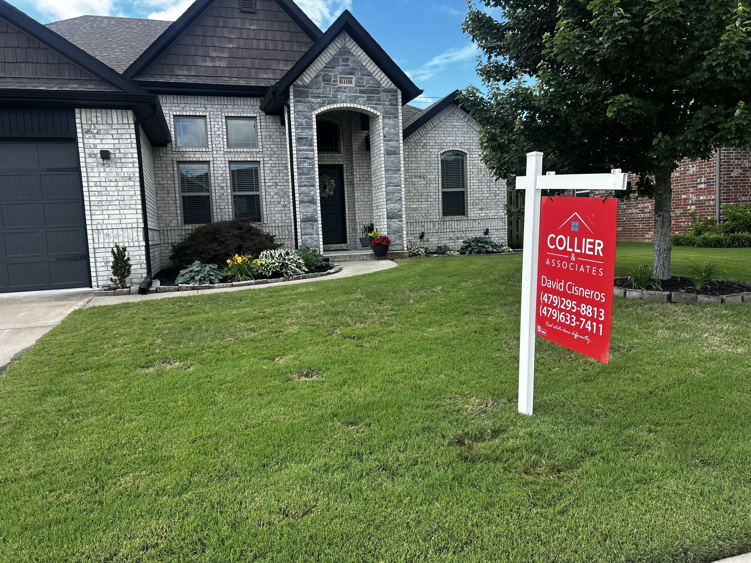 A modern brick house with a grey front door and a driveway. There is a red real estate sign in the front yard with contact information for a real estate agent named David Cisneros. The yard has green grass, small bushes, and a large tree on the right.